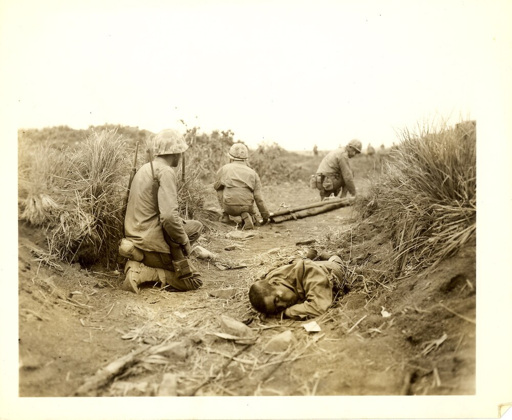 A Marine kneels next to a fallen Marine while stretcher bearers wait for orders to move to a Marine they can help to save.