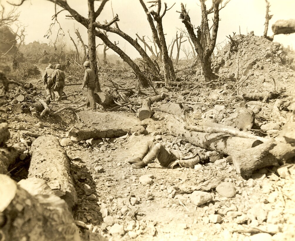 A dead Japanese soldier in foreground and three Marines advancing forward.