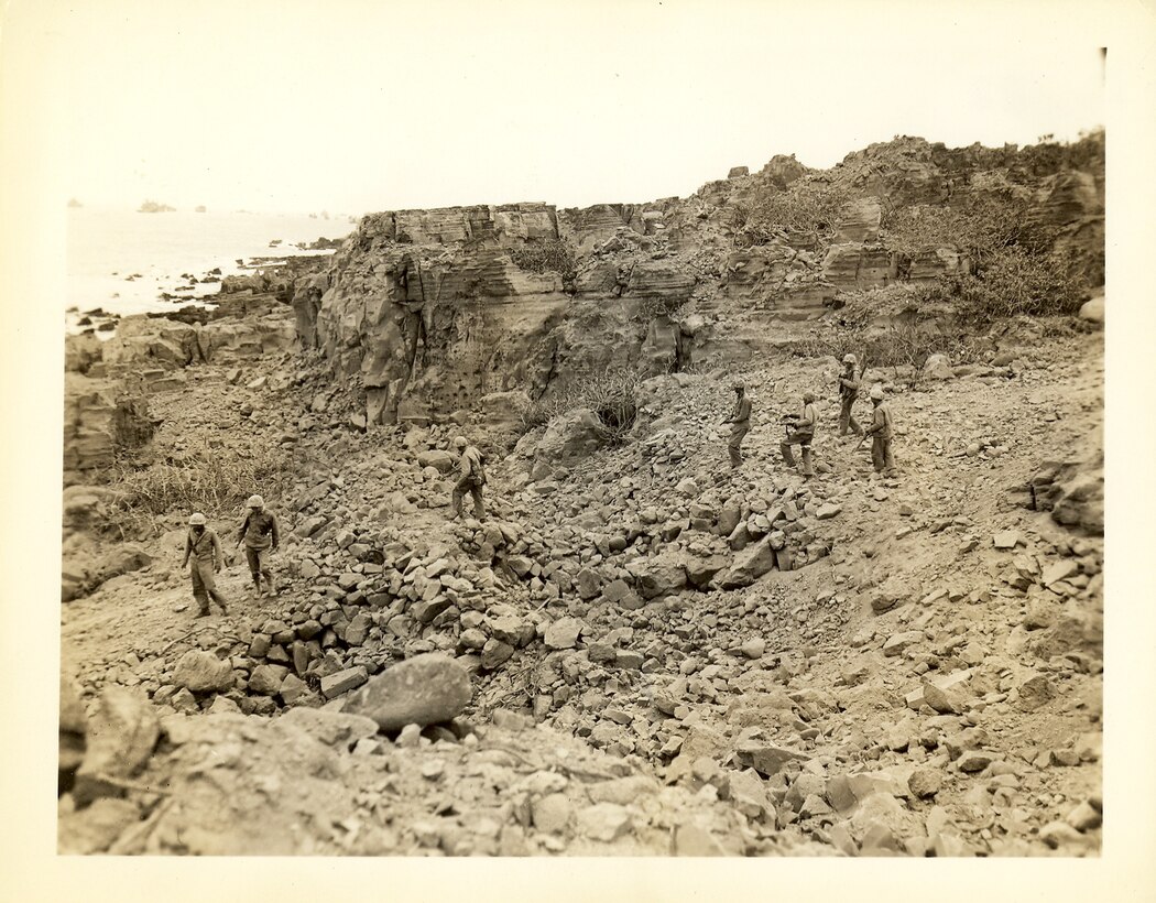 Marines patrol rocky terrain, U.S. Fleet is staged off shore in background.