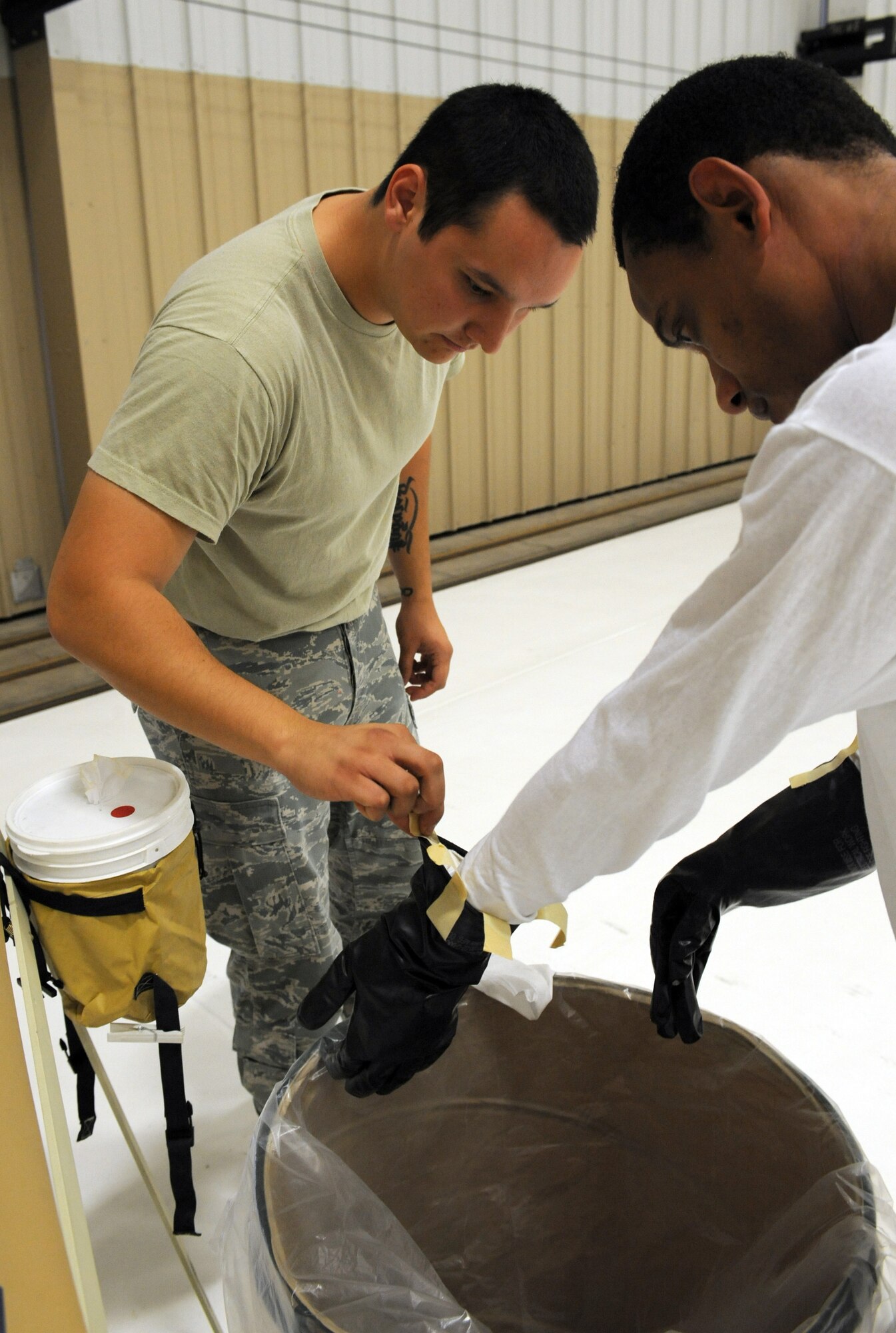 U.S. Air Force Senior Airman Jose Ibarra, 57th Weapons Support Squadron, Nellis Air Force Base, Nev., helps Staff Sgt. Kendrick Walker, 7th Operations Support Squadron, Dyess AFB, Texas, remove his gloves during aircrew decontamination training on Davis-Monthan AFB, Ariz., Sept. 21, 2012. The mitigation process is setup into two separate areas: a "clean" and "dirty" side. This ensures that personnel working on the dirty side do not bring contaminates to the clean side. It also allows personnel working on the clean side to easily replace workers on the dirty side in case of a mishap. (U.S. Air Force photo by Airman 1st Class Timothy Moore/Released)