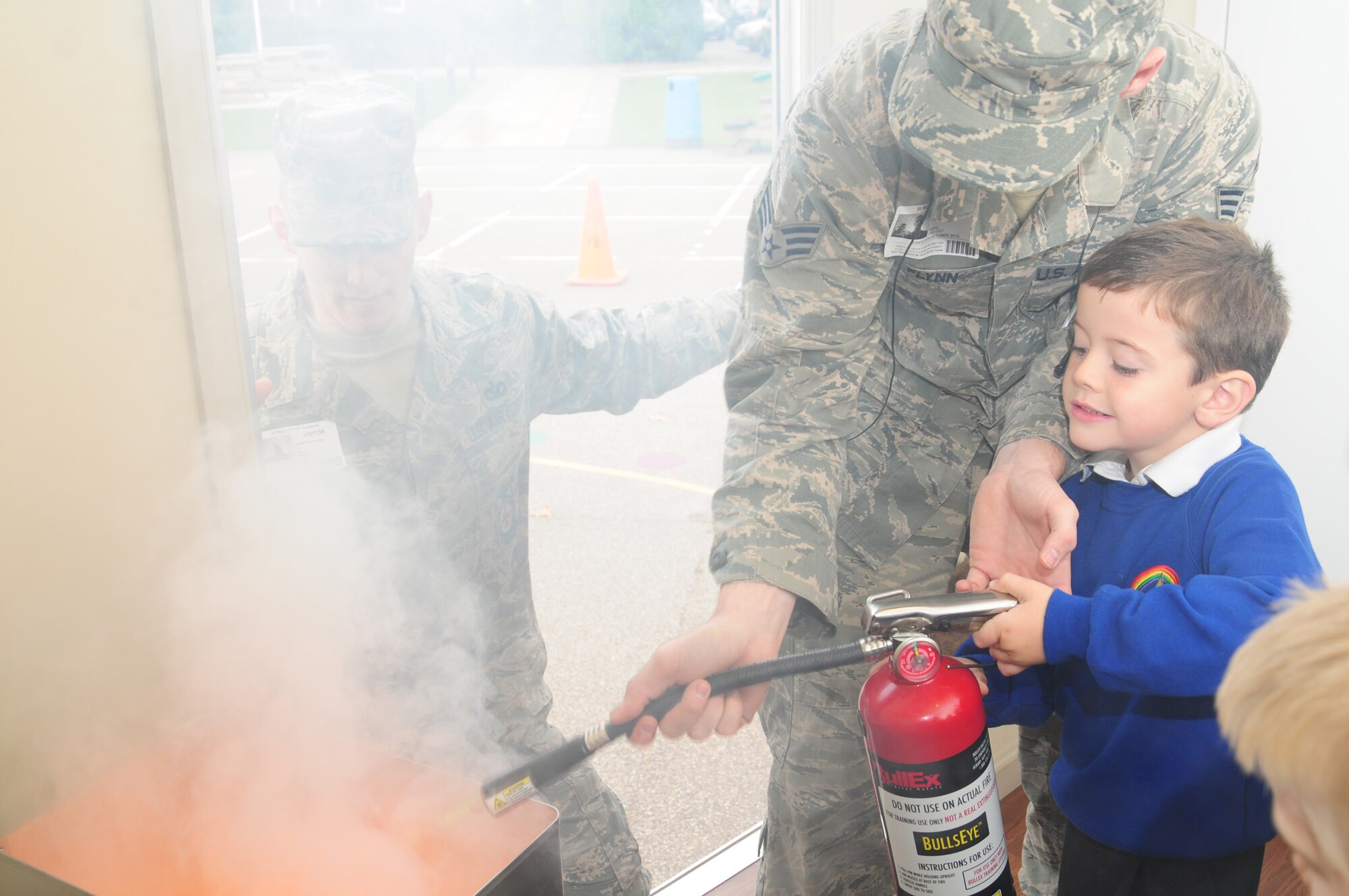 Senior Airman Patrick Flynn, center, helps Jack Gathercole, 5, put out a simulated fire in the Fire Prevention trailer Oct. 8, 2012, at St. Mary's Academy in Mildenhall, England, as Staff Sgt. Andrew Cranfill looks on. Airmen, all firefighters from RAF Mildenhall Fire Department, and Sparky the Fire Dog, visited the school during Fire Prevention Week. (U.S. Air Force photo/Karen Abeyasekere)