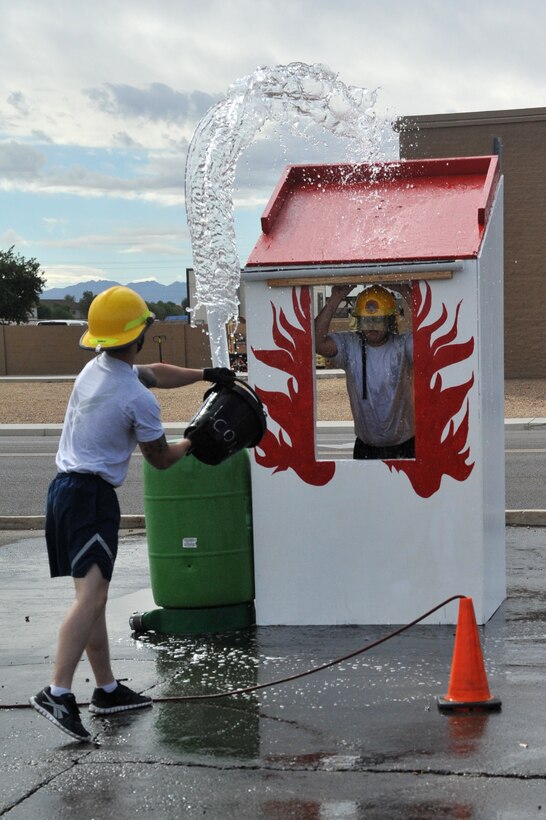 Tech. Sgt. Robert Simpson, 56th Civil Engineer Squadron pavements and heavy equipment operator, pretending to be a victim of a burning house, watches Senior Airman Jason Reed, 56th CES pavements and heave equipment operator, as he tosses water on the roof during the Bucket Brigade obstacle of the 2012 Firefighter’s Muster Oct. 12 at Luke Air Force Base, Ariz. (U.S. Air Force photo by Senior Airman Sandra Welch)