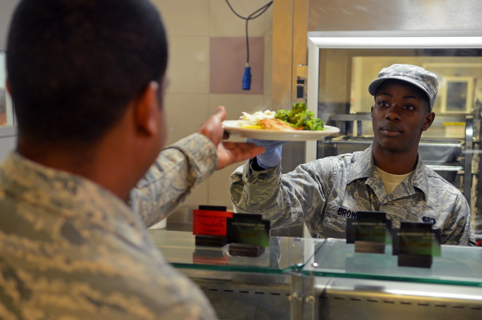 Airman 1st Class Jemodric Brown, 31st Force Support Squadron services journeyman, hands a plate of food to an Airman at the La Dolce Vita Dining Facility at Aviano Air Base, Italy, Oct. 24. The menu at the dining facilities is not changing, but the way food is prepared and cooked is. Green food items are cooked in a healthier manner, such as grilling chicken instead of frying it. (U.S. Air Force photo/Airman 1st Class Matthew Lotz)