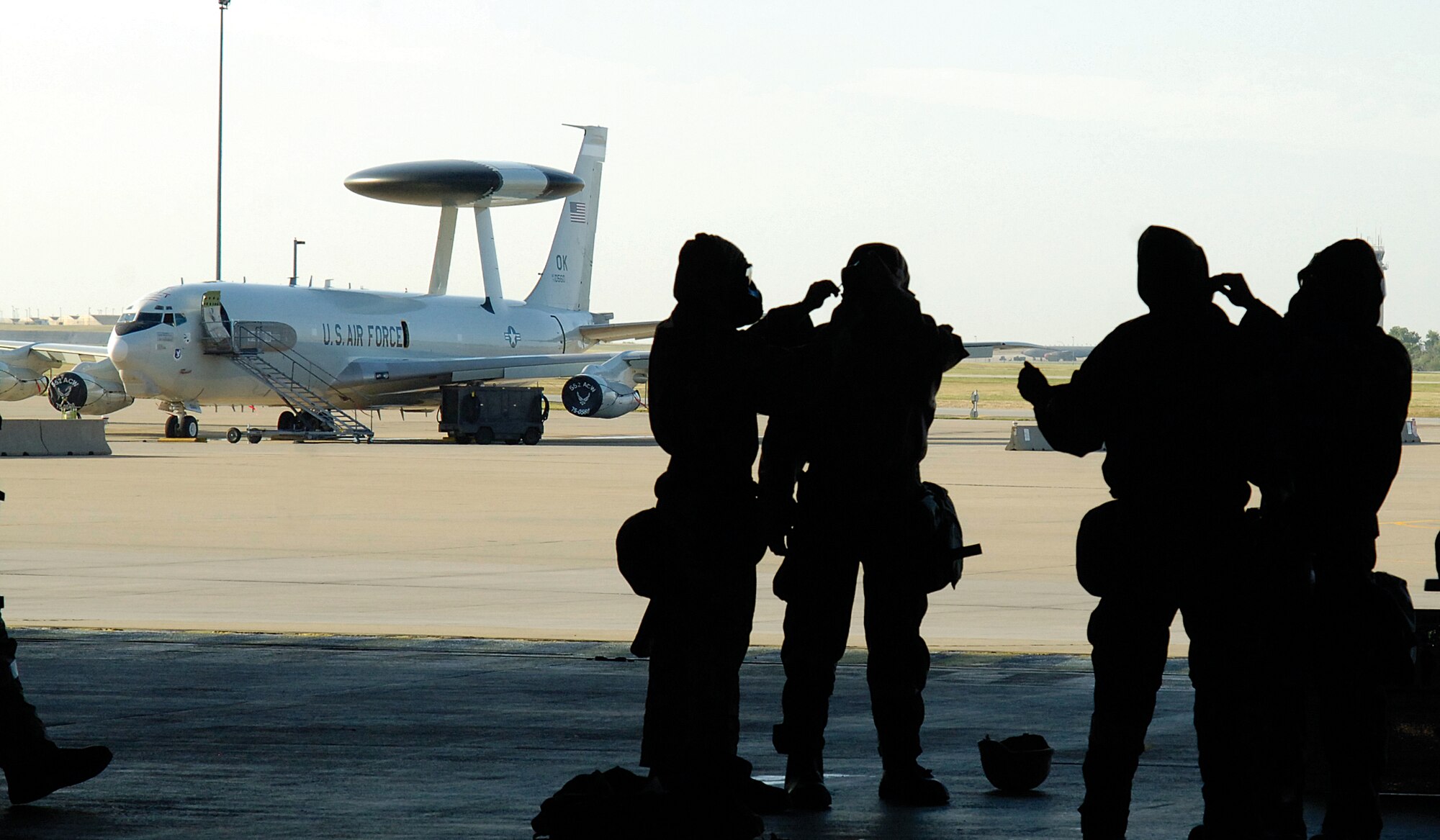 Airmen help each other correctly put on the protective uniform, MOPP gear, in a training station just yards from an E-3 Sentry aircraft parked on the ramp. (Air Force photo by Margo Wright)