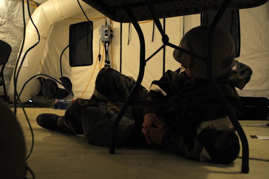 A U.S. Air Force Airman assigned to the 4th Fighter Wing lays under a table in full battle ensemble during increased alarm and Mission Oriented Protective Posture conditions at Base X on Seymour Johnson Air Force Base, N.C., Sept. 20, 2012. Military personnel must don their chemical protective overgarment and helmets and take cover in a hardened shelter during increased alarm and MOPP conditions. (U.S. Air Force photo/Airman 1st Class Aubrey Robinson/Released)