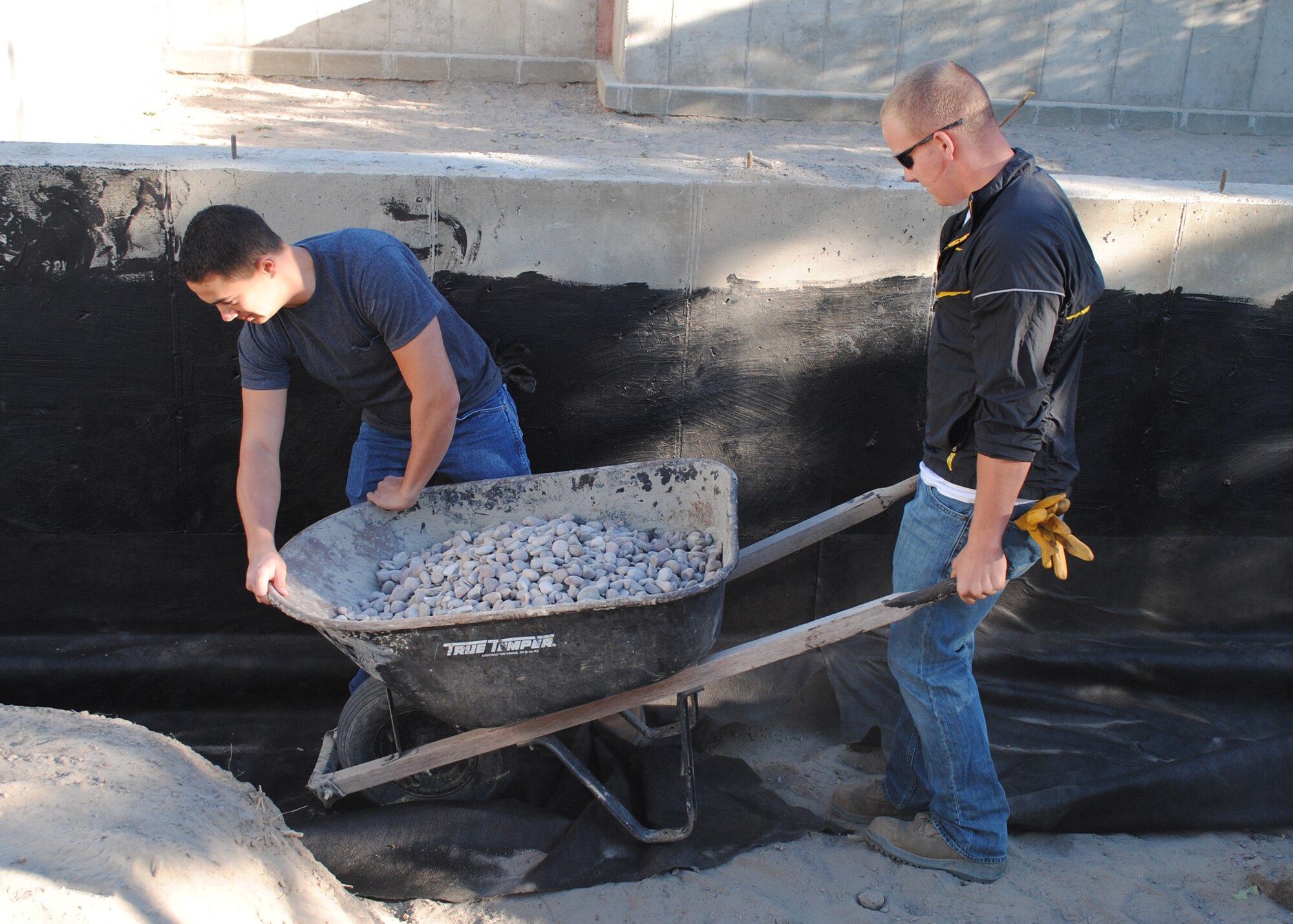 (From left) Airmen 1st Class Juan Rigionimolina, and Dalton Mays, both 819th RED HORSE Squadron members, prepare to carefully pour gravel over a pipe during a Habitat for Humanity housing project. Organized by the Big Sky Top 3 Association, the all-day project had a crew of 15 to 20 Airmen from the 341st Logistics Readiness Squadron and the 819th RHS shoveling gravel, digging, and installing a French drain and a pipe to prevent water from seeping through the foundation. (U.S. Air Force photo/Airman 1st Class Katrina Heikkinen)