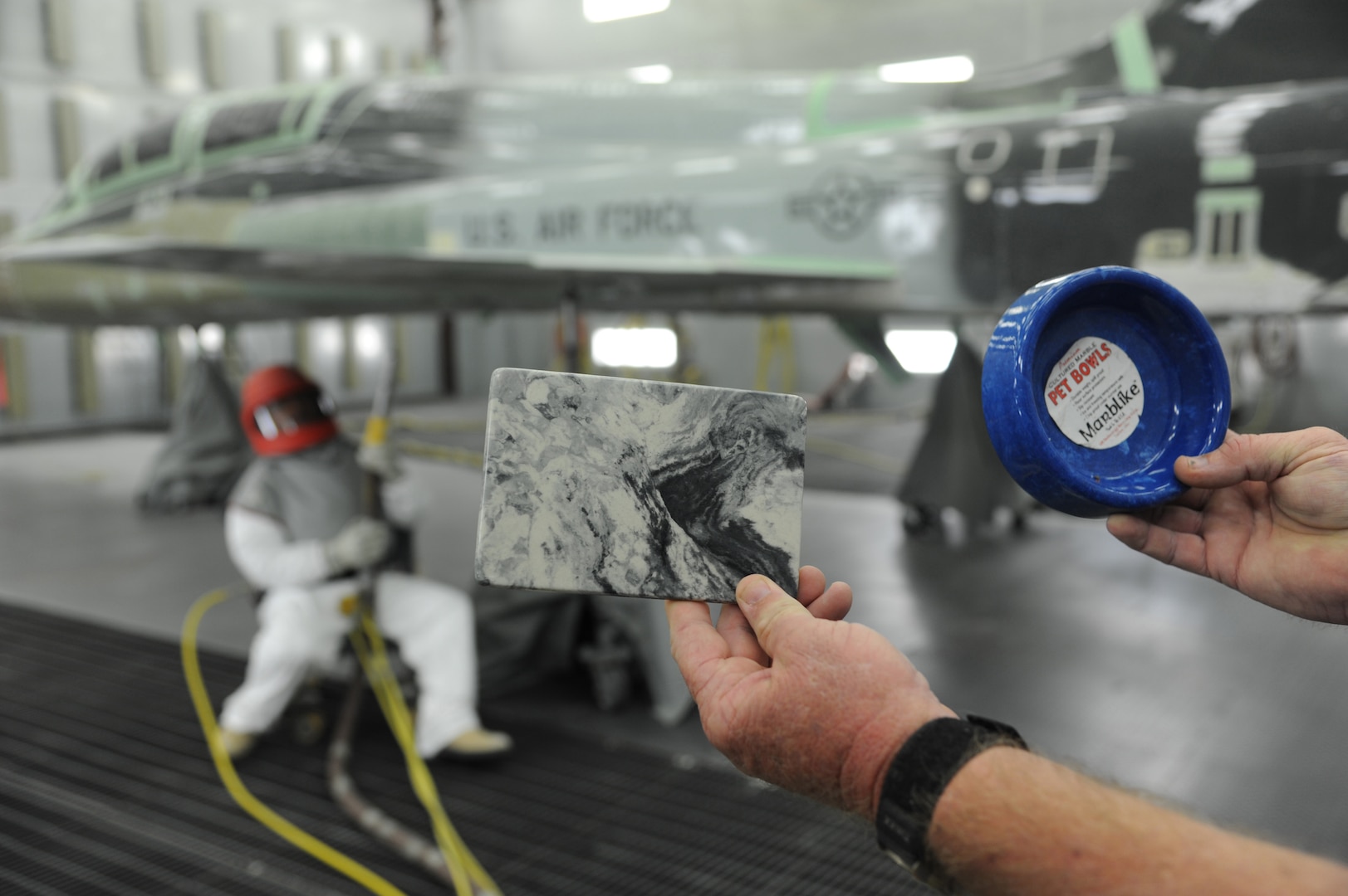 John Harris, 12th Flying Training Wing Maintenance Division media blast technician, holds sample items made from recycled media blast material as John Esquivel (left) uses corn starch to strip the paint from a T-38 Talon at Joint Base San Antonio-Randolph, Sept. 20. Two materials 12th FTW shops air blast to remove paint from T-1, T-6, T-38 and F-16 aircraft are plastic and corn starch, which have been deemed environmentally safe by the Air Force.
Randolph generates about 20,000 pounds of plastic and 28,500 pounds of corn starch annually that is recycled.  The excess plastic is shipped to Canton, Ohio, where it is manufactured into products such as simulated marble tiles and dog bowls.  (U.S. Air Force photo by Rich McFadden) 