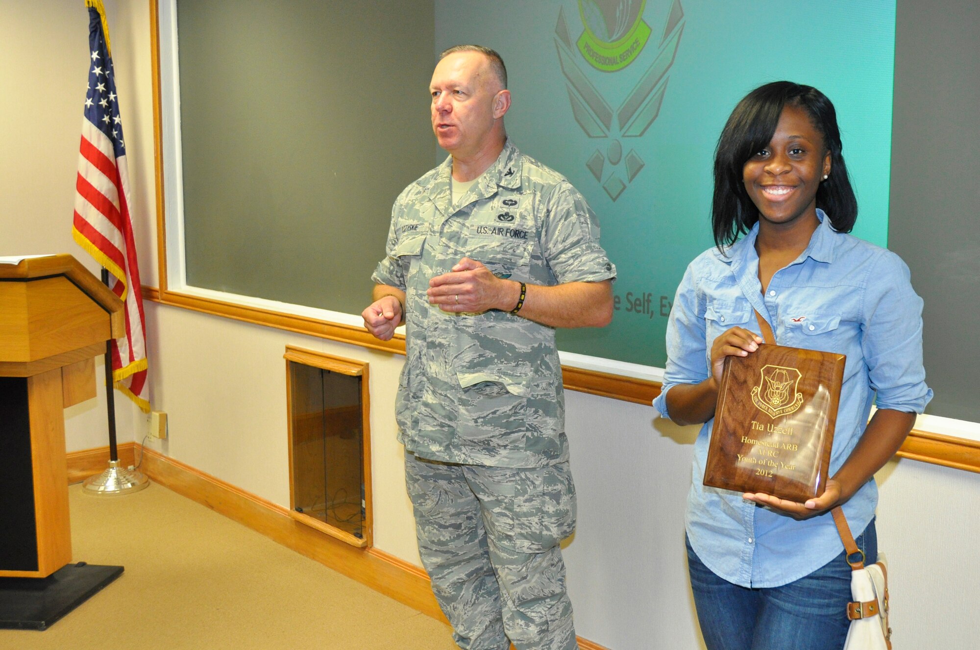 Ms. Tia Uzzell holds up her plaque for winning the 2012 Homestead Air Reserve Base Air Force Reserve Command Youth of the Year Award, Aug. 3.  Col. Elwin Rozyskie, 482nd Mission Support Group commander, list her accomplishments in the background during the Unit Training Assembly. (U.S. Air Force photo/ Tech. Sgt. Lou Burton)