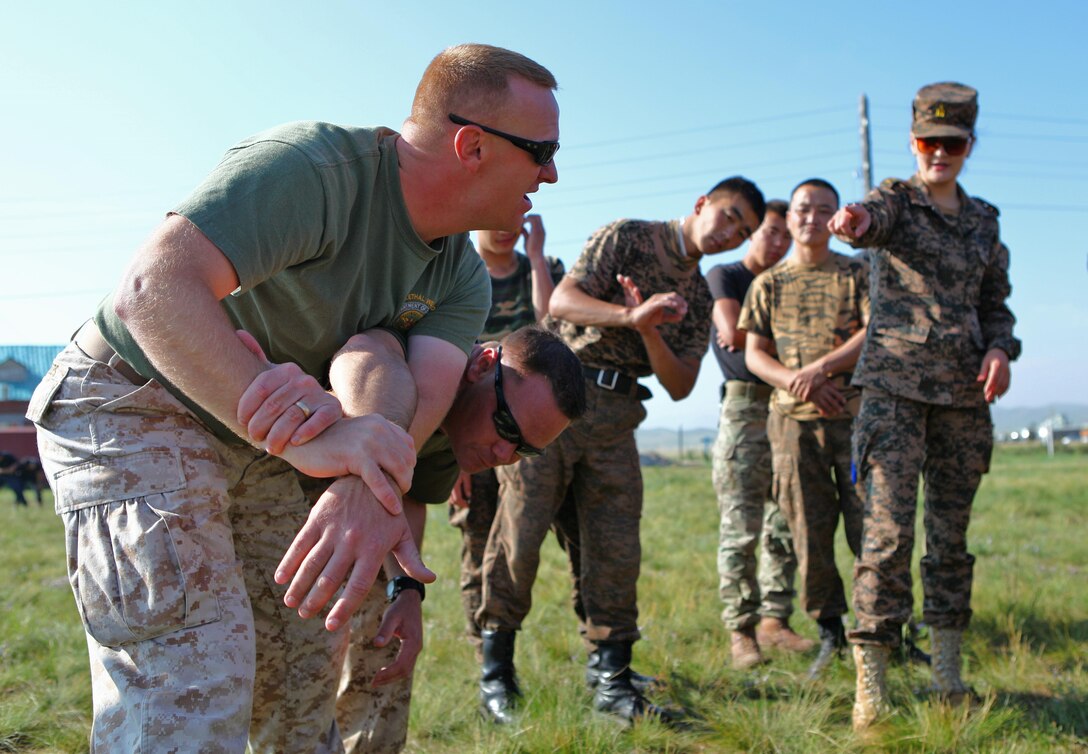 Staff Sgt. Alan Stowers (left) and Gunnery Sgt. Mark Massalski demonstrate Mechanical Advantage Control Hold (MACH) techniques as Mongolian Armed Forces Sgt. L. Bolormaa (right), an instructor with the MAF's 330 Peacekeeping Battalion, provides translation during the field training portion of the Non-Lethal Weapons Executive Seminar at Five Hills Training Area, Mongolia, Aug. 18. Mongolian troops and police personnel are rehearsing MACHs to further develop non-lethal control techniques for use during low-intensity conflict situations. The training is taking place as part of Non-Lethal Weapons Executive Seminar (NOLES) 2013, a regularly scheduled field training exercise and leadership seminar sponsored by U.S. Marine Corps Forces Pacific, and hosted annually by various nations throughout Asia-Pacific. In its 12th iteration, this multinational training event is designed to promote awareness and effective use of non-lethal weapons. Mongolia, a regular participant, is hosting NOLES for the third time. Stowers, a platoon sergeant an non-lethal weapons instructor with Law Enforcement Battalion, 3rd Marine Expeditionary Force Headquarters Group, III MEF, is from Garber, Okla. Massalski, also a platoon sergeant and instructor with LE Battalion, is from Townsend, Mass.