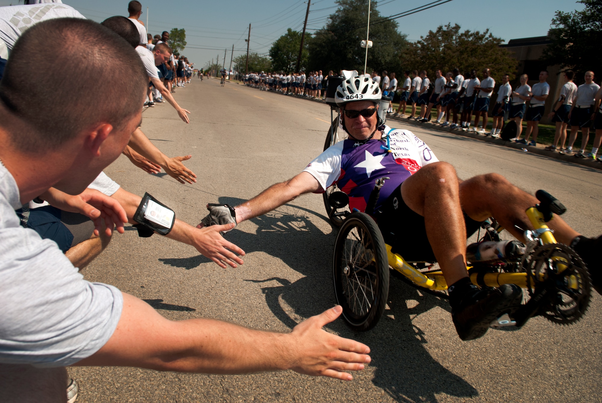 A Hotter'N Hell Hundred rider greets Airmen in training at Sheppard Air Force Base, Texas, on his way out of the base Aug. 24, 2013. After "Airpower Alley" and a rest stop, bicycle riders rode through a gauntlet of hundreds of Airmen screaming and cheering them on to the finish line. The annual race is in its 32 year and celebrates the founding members of the Texoma area with a 100-mile race in 100-degree temperatures. (U.S. Air Force photo/Staff Sgt. Mike Meares)
