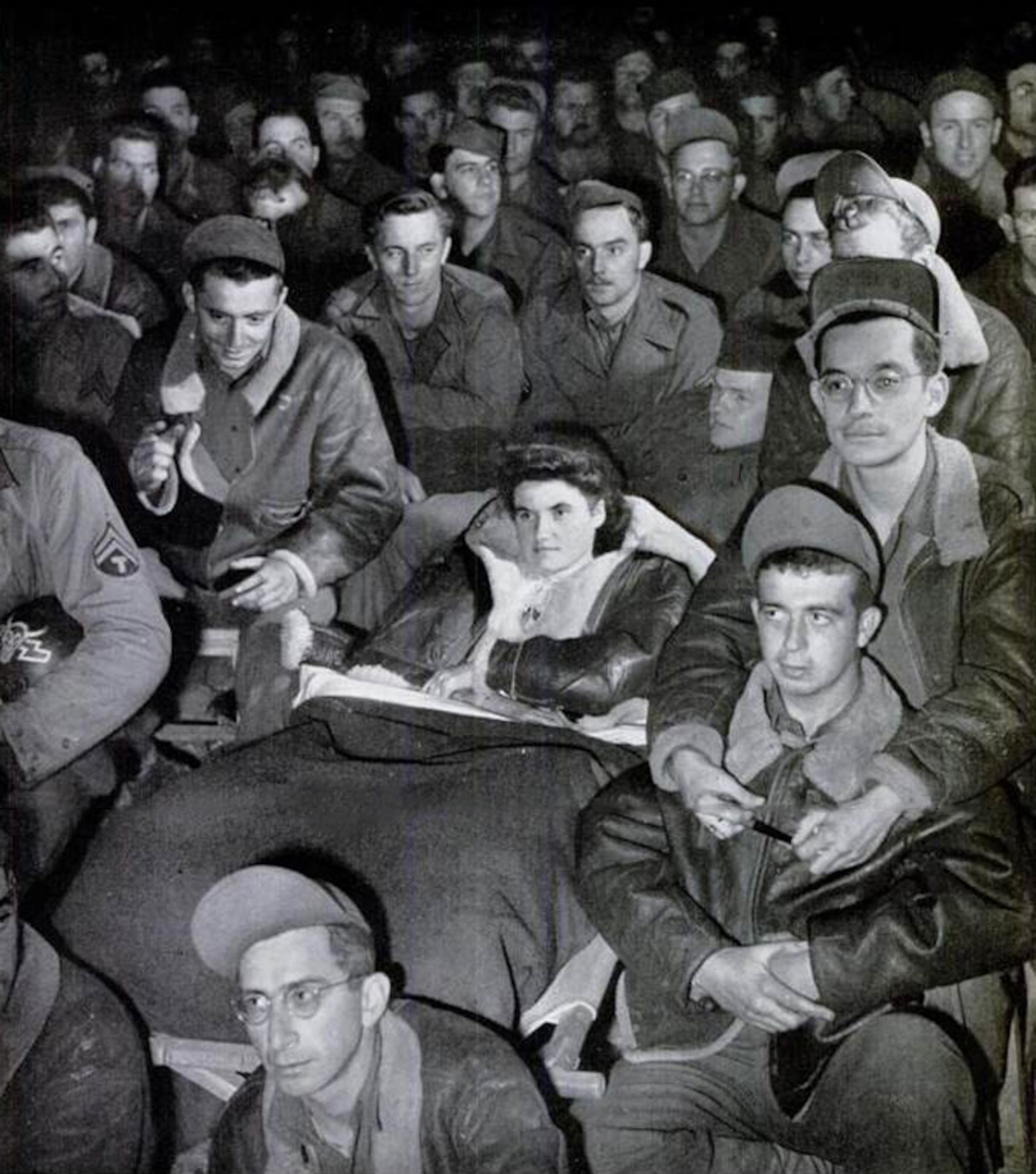 Yvette Hamel takes in some entertainment with the men of the 371st Fighter Group at an airfield in France.  This photograph was published on page 45 of the November 27, 1944, issue of LIFE Magazine.  The caption for this photo was:   “LEGLESS YVETTE HAMEL, DRESSED IN A FLYING JACKET AND BLOUSE MADE OF PARACHUTE SILK, WATCHED A GI SHOW WITH MEN OF THE FIGHTER GROUP WHICH ADOPTED HER.”

The short article beneath LIFE’s photo included the following details of Yvette’s remarkable story:

AIRMEN’S WARD
U.S. fliers in France adopt girl who lost both legs in Normandy

Last July 16-year old Yvette Hamel was milking a cow in a field near the Norman town of La Haye du Puits.  A German shell landed in the field, wounding her so badly that both of her legs had to be amputated.  A few days later, Colonel Bingham Kleine, commanding officer of a 9th AF fighter group, met her in a near-by hospital.  Colonel Kleine saw that she was moved to a better hospital, finally arranged to have her treated in his group dispensary.  When the group moved, Yvette moved with it, cared for by several hundred men and two French Red Cross volunteers.

The men of Colonel Kleine’s group are taking fine care of Yvette. Among themselves they have raised more than $2,000 for new hospital bills and have spared no trouble to keep her comfortable.  In the midst of all this attention Yvette is cheerful, although she cries a little when she thinks no one is around.  She doesn’t speak much English but is learning the curious GI brand as quickly as anyone. (LIFE Magazine)
