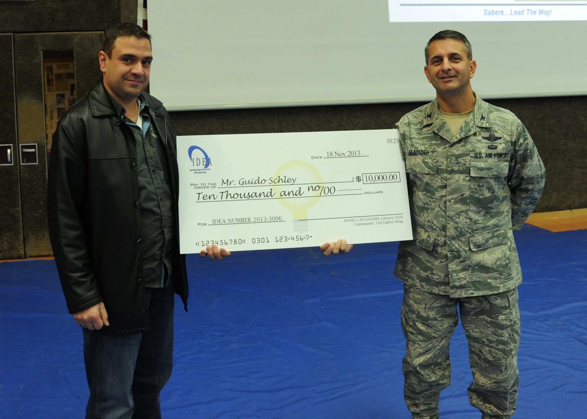 SPANGDAHLEM AIR BASE, Germany --- Col. David Julazadeh, commander of 52nd Fighter Wing, right, presents Guido Schley, a 52nd Civil Engineer Squadron Heating, Venting and Air Conditioning foreman of Dodenburg, Germany, with a large check before more than 1,000 Saber Airmen during a commander’s call at the Skelton Memorial Fitness Center Nov. 18, 2013. Schley earned the check for his submission through the Innovative Development through Employee Awareness program. The IDEA program is an incentive system where nominated applicants may receive cash awards for their approved proposals to enhance processes or improve productivity and efficiency that benefit the Air Force and government.  (U.S. Air Force photo by Airman 1st Class Dylan Nuckolls / RELEASED)