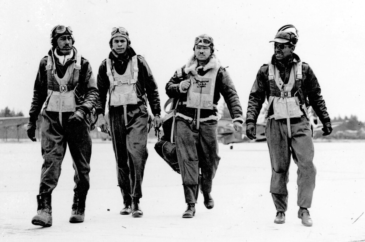 Four Tuskegee airmen walk on a flight line in black and white photo.