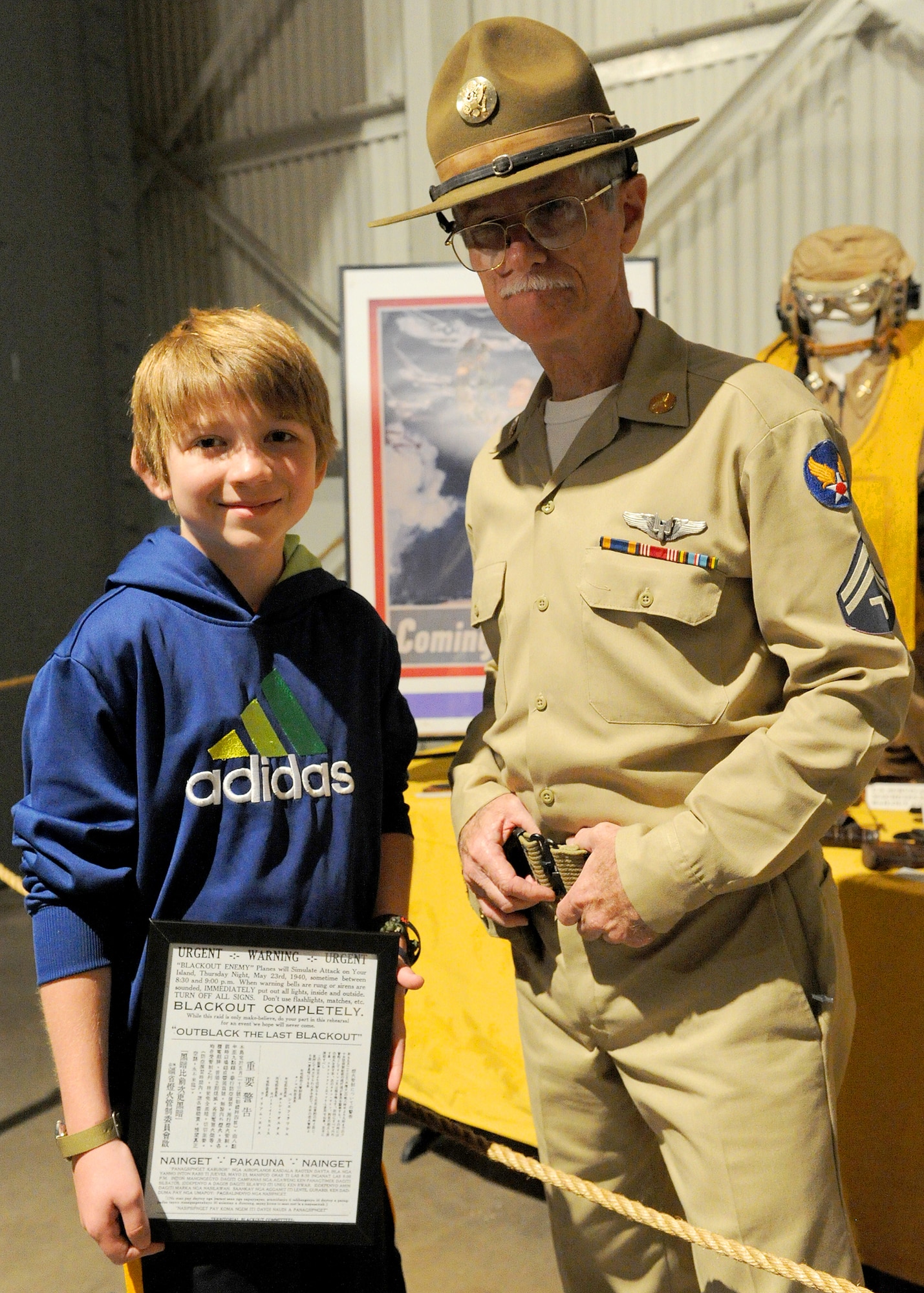 Carl Hess, Make-A-Wish recipient, poses for a photo while holding a blackout restriction certificate with Army Aviator (retired) Jerryl Stanfield at the Pacific Aviation Museum, Ford Island, Hawaii, Feb. 19, 2013. A blackout during war, or in preparation for an expected war, is the practice of collectively minimizing outdoor light, including upwardly directed (or reflected) light. This was done in the 20th century to prevent crews of enemy aircraft from being able to navigate to their targets simply by sight. Carl is fighting acute lymphoblastic leukemia (ALL), a fast-growing cancer of the white blood cells, and wished to be a downed World War II pilot. During his visit he received a rare opportunity to spend the night aboard the USS Missouri, fly in a bi-plane, and tour various historical WWII sites. (U.S. Air Force photo/Tech. Sgt. Jerome S. Tayborn)
