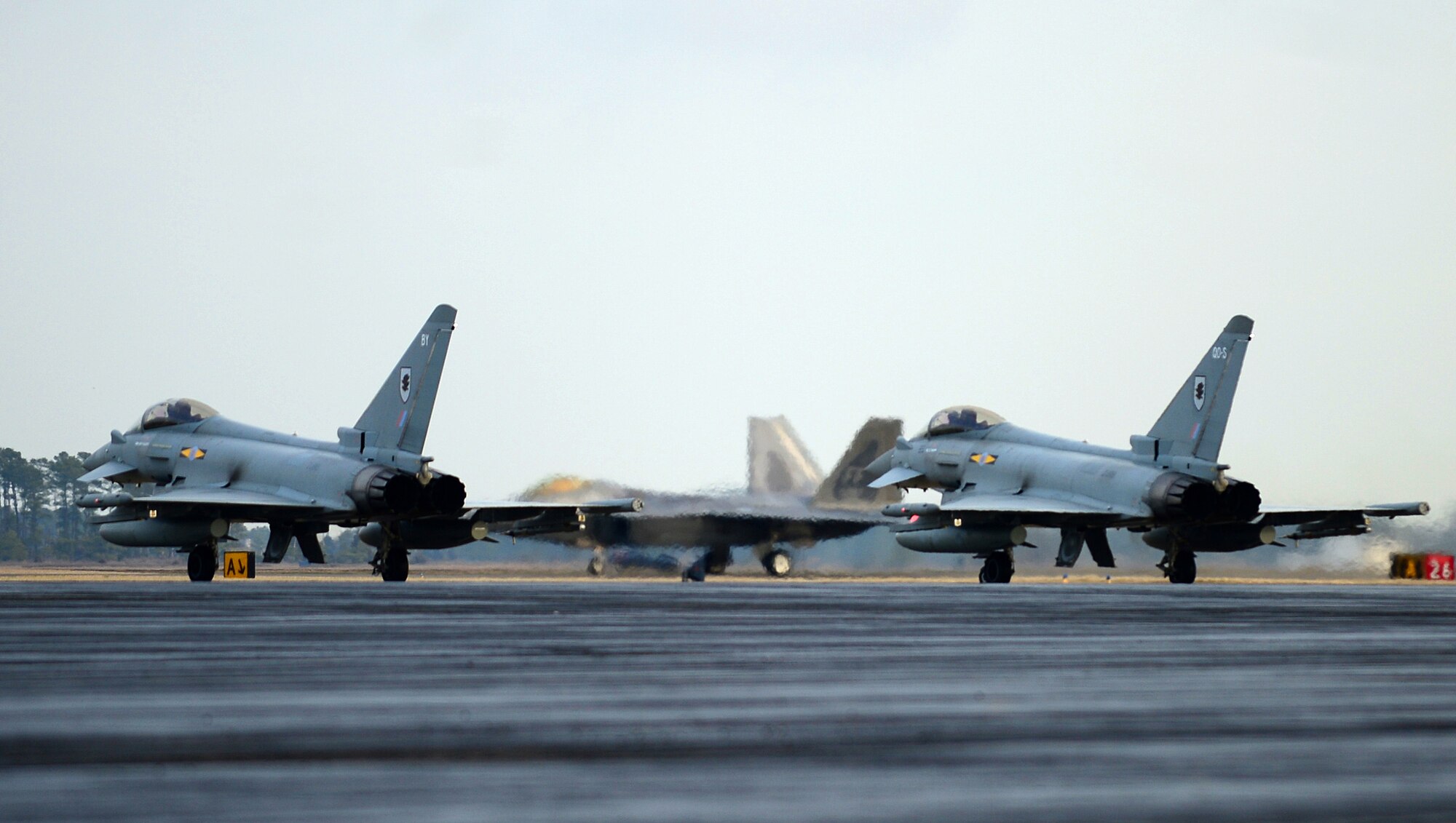 Two Typhoon FGR4s wait as an F-22 Raptor prepares to take off at Langley Air Force Base, Va., Jan. 30, 2013. In order to enhance the joint capability of the Typhoon FGR4, XI Squadron RAF is operating with the F-22 Raptor.  (U.S. Air Force photo by Senior Airman Kayla Newman/Released)