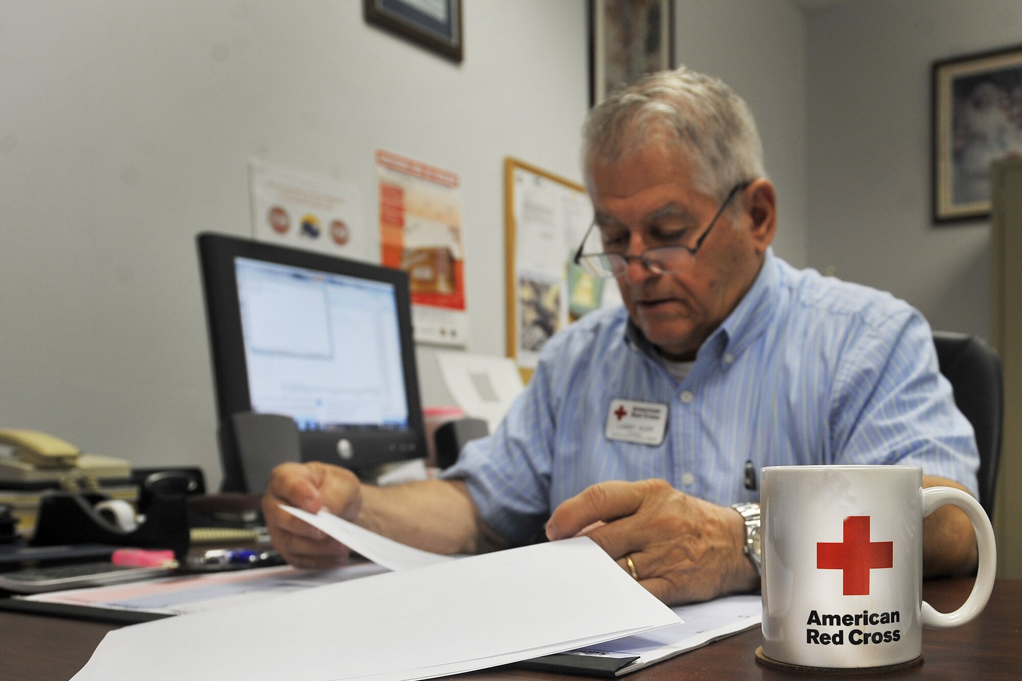 An American Red Cross mug sits in front of Larry Huff, 20th Medical Support Squadron station chairman, as he sits in front of his desk while going through his training procedures, Shaw Air Force Base, S.C., July 16, 2013. Huff has been involved with the Red Cross since 2004. (U.S. Air Force photo by Airman 1st Class Ashley L. Gardner/Released)