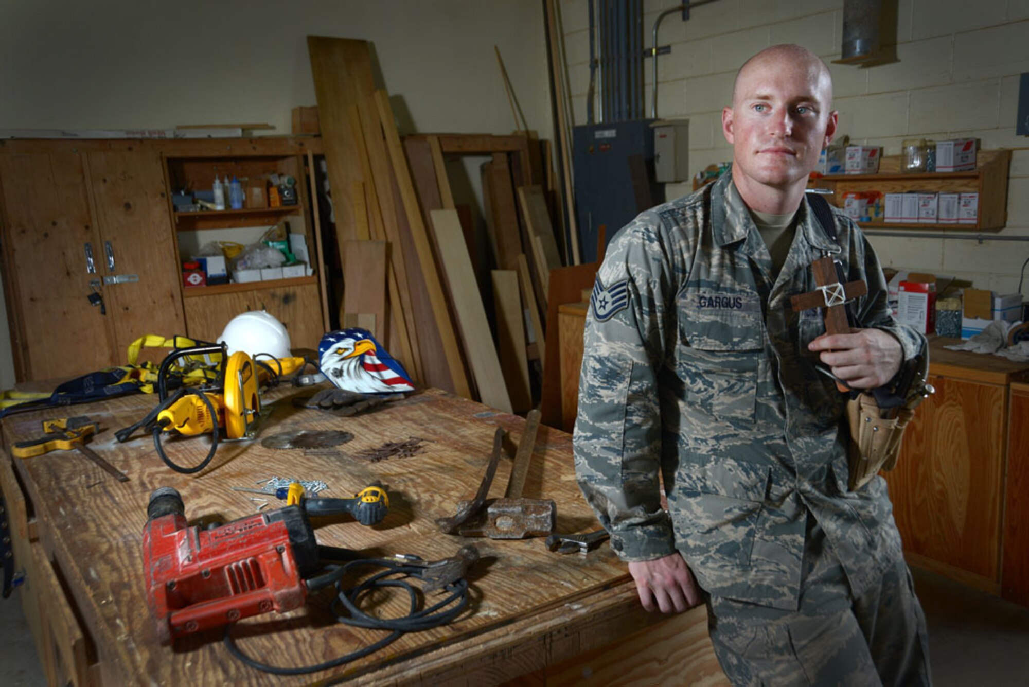U.S. Air Force Staff Sgt. Eric Gargus, 18th Civil Engineer Squadron structural craftsman, stands surrounded by the tools of his trade while holding a cross that he crafted in his free time. Gargus recently returned from an impact trip to Thailand where he helped build a playground for children who live in homes supported by the organization Remember Nhu. (U.S. Air Force photo by Tech. Sgt. Jocelyn L. Rich-Pendracki/Released)