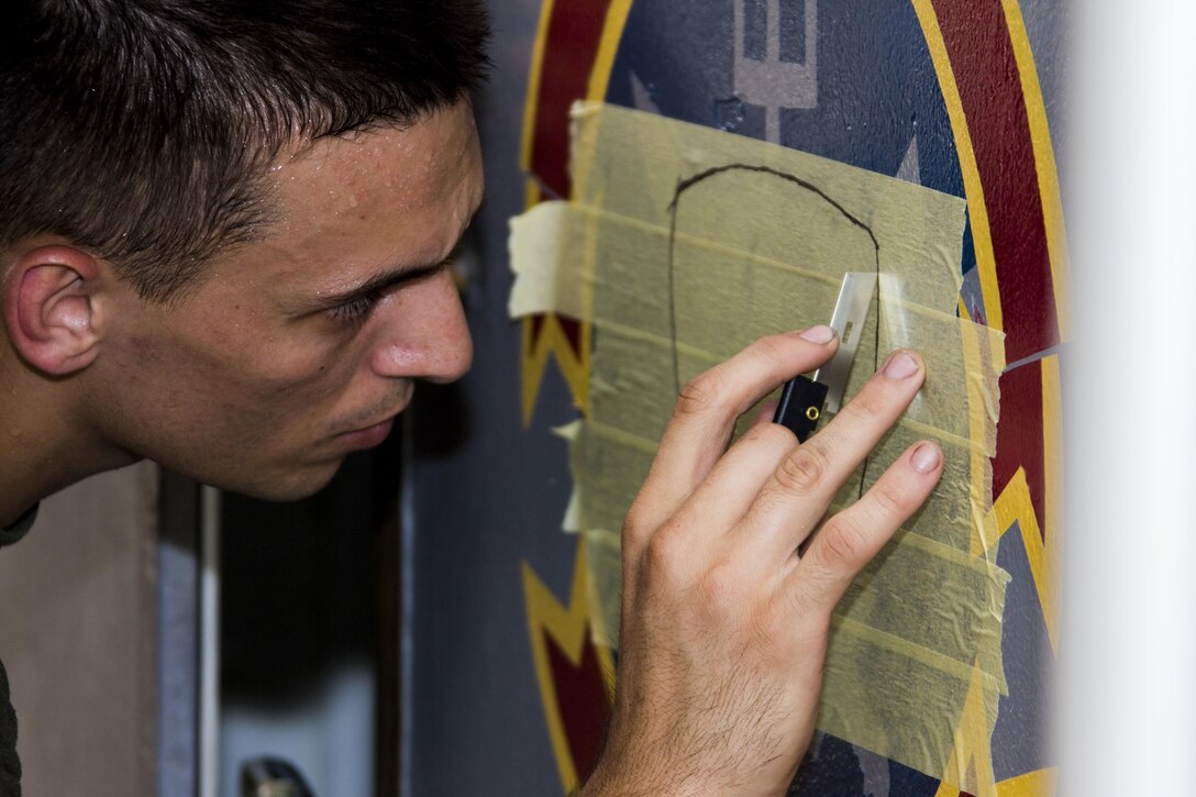 U.S. Marine Corps Cpl. Nicolas Belmer with Marine Medium Tiltrotor Squadron (VMM) 266 (Reinforced), 26th Marine Expeditionary Unit (MEU), cuts tape while painting the 26th MEU emblem incorporating a rooster onto double-wide doors on the flight deck of the USS Kearsarge (LHD 3), at sea, July 26, 2013. The 26th MEU is a Marine Air-Ground Task Force forward-deployed to the U.S. 5th Fleet area of responsibility aboard the Kearsarge Amphibious Ready Group serving as a sea-based, expeditionary crisis response force capable of conducting amphibious operations across the full range of military operations. (U.S. Marine Corps photo by Cpl. Kyle N. Runnels/Released)
