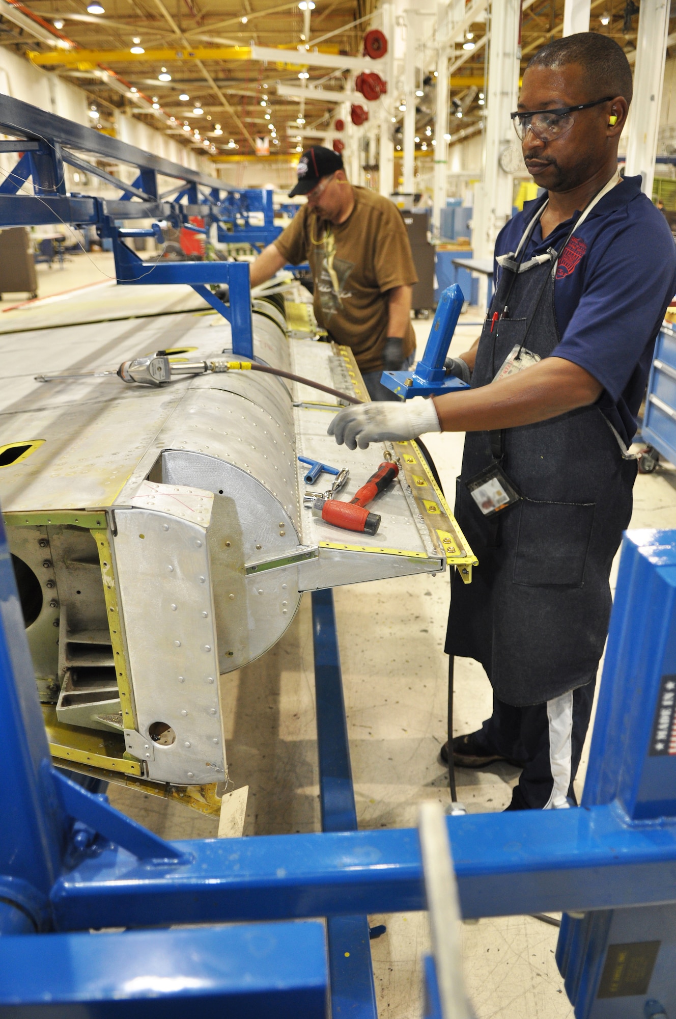 Demerick Clifton, front, and Nathanuel Cloud, sheet metal mechanics with the KC-135/E-3 rudder repair shop, work on replacing rudder bearings while the rudder is braced in a fixture. (Air Force photo by Micah Garbarino)