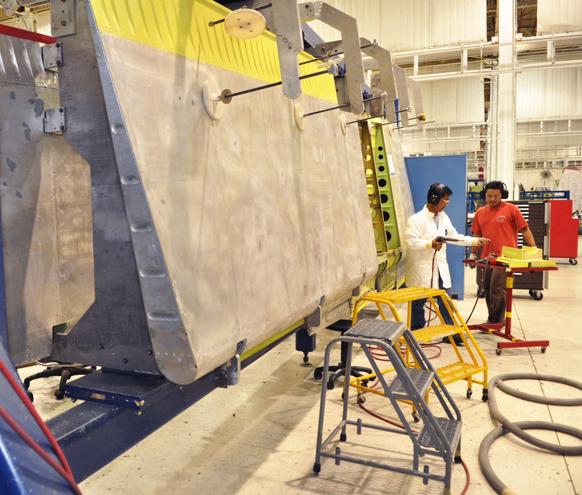 Elton Nguyen, left, and Thien Le ready the PCU fitting before placing it on the rudder. The rudder is suspended in a brace while the work is performed. Cranes, suspended from overhead beams, and dollies move the rudders from one work station to the next. Both Mr. Nguyen and Mr. Le work in the 76th Commodities Maintenance Group. (Air Force photo by Micah Garbarino)