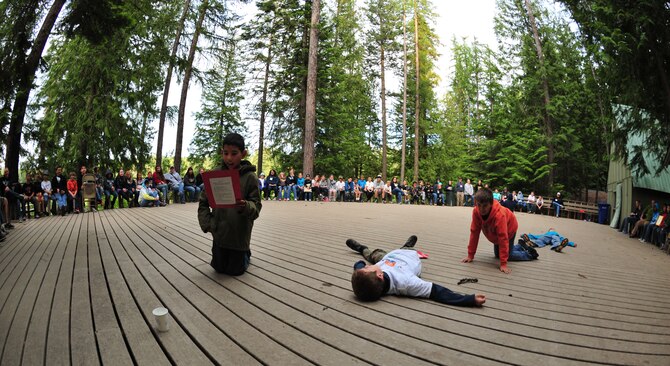 (Left to Right) Fourth graders Andrew, Elliott, Dominic and Sawyer, also known as their camp name Kinunka, act out a skit as teachers, camp counselors and other fourth grade students look on May, 30, 2013, during Michael Anderson Elementary School’s annual camping trip to YMCA Camp Reed. For this particular skit, the boys had to pretend they were stranded in a desert, and out of nowhere, a cup of water appears as they struggle to reach it. (U.S. Air Force photo by Senior Airman Earlandez Young/Released)   