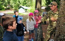 Airman 1st Class Samuel Wallace, Survival, Evasion, Resistance and Escape specialists, demonstrates to Michael Anderson fourth graders how to quickly build a place for shelter in the wilderness using a poncho and tying it to trees and other steady objects in the forest May 30, 2013, at YMCA Camp Reed. This was a part of the wilderness survival session at the school’s annual Camp Reed camping trip. Students learned other survival techniques such as what insects are safe to eat, how to camouflage by using face paint and more. (U.S. Air Force photo by Senior Airman Earlandez Young/Released)   