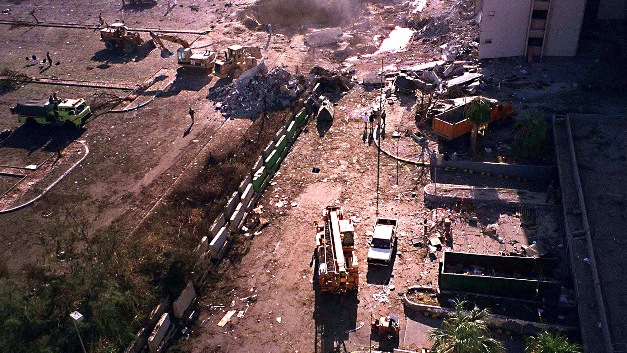 The crater caused by the explosion of a fuel truck outside Khobar Towers on Tuesday, June 25, 1996, is visible at the top of this photograph of the facility taken from a nearby building on King Abdul Aziz Air Base near Dhahran, Saudi Arabia, on June 26, 1996. The dotted line running diagonally through the photo is the concrete barrier, sometimes known as a Jersey Wall, which is credited for deflecting the blast of the explosion upward and reducing its effect. (DoD photo/Released)