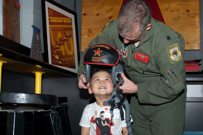 Lt. Col. Michael Shepherd, 64th Aggressor Squadron assistant director of operations for academics, helps Ryan Navarrate-Pak, a childhood cancer survivor, put on a flight helmet  in the 64th AGRS heritage room June 8 2013, at Nellis Air Force Base, Nev. Navarrate-Pak and his family were treated to a tour of Nellis by Airmen from the 64th AGRS with help from the 65th AGRS, the 547th Intelligence Squadron, the United States Air Force Air Demonstration Squadron Thunderbirds and the 99th Civil engineer squadron. (U.S. Air Force Photo by Staff Sgt. Gregory Brook)