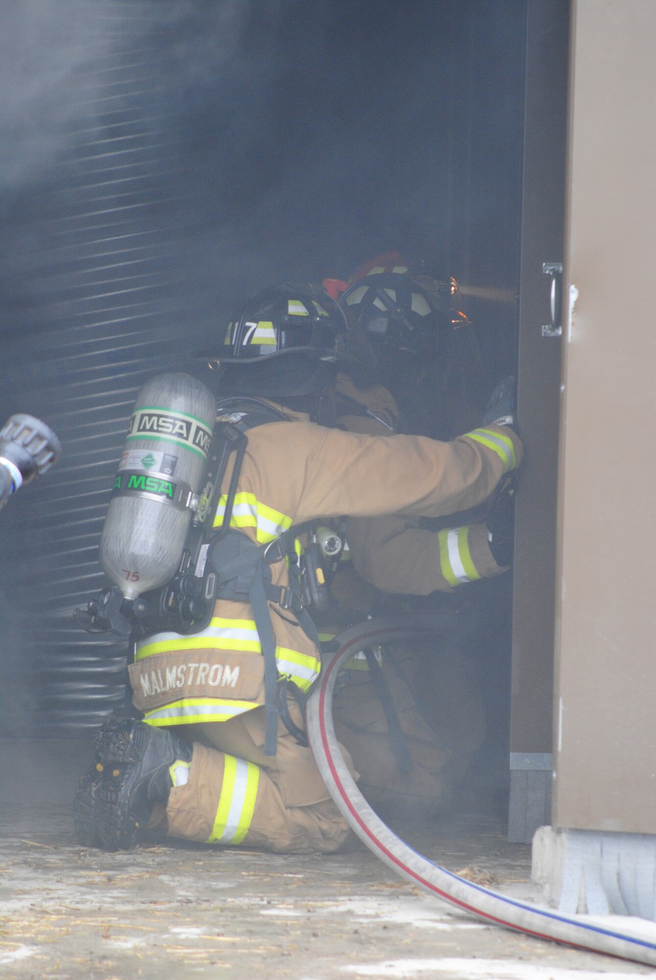 Two Air Force Academy cadets touring Malmstrom’s fire department, alongside one of Malmstrom’s firefighters prepare to enter a room in the fire trainer that holds a live fire.  The cadets toured the training facility, received safety briefings and were able to handle the fire hoses before participating in the exercise.  (U.S. Air Force photo/Senior Airman Cortney Paxton)