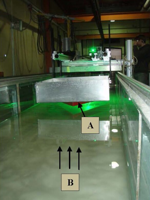 A New York University doctorate student monitors the laser in ERDC Cold Regions Research and Engineering Laboratory’s flume recently, looking at the sea ice and ocean interface.  The “V” is attached to the underside of the bow, as indicated by (A) in the photo. The “V” deflects the air bubbles, allowing the laser to track the glass spheres’ circulation in the water current. The flow direction is indicated by the arrows at (B) and also shows the rippling effect of the scalloped ice.