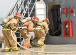 Crewmembers prepare to fight a simulated fire onboard the LCU 2001”Runnymede.” If a fire were to break out, half of the fire team would immediately start spraying down the area of fire while the other half dons fireproof suits. On  half the team is dressed, they switch off positions so they can get dressed while still maintaining constant control of the fire. The crew is expected to take no more than two minutes to be fully dressed in fire protection gear.