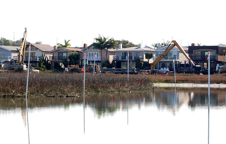 White poles mark the edge of the channels to indicate areas from which CJW will dredge material. Red flags indicate sensitive marsh areas for heavy equipment to avoid. Equipment in the background is in the process of removing material and placing it into trucks for transportation to upland disposal sites. 