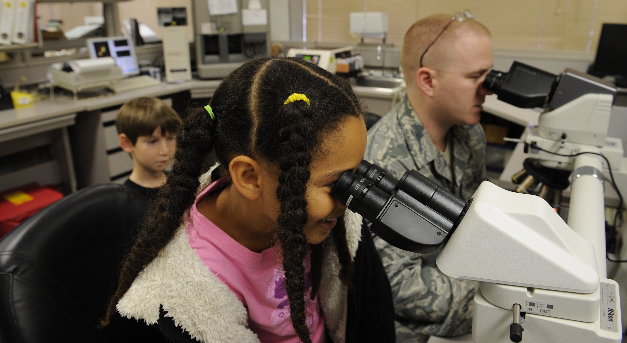 A Stearley Heights Elementary student peers through a microscope as U.S. Air Force Staff Sgt. Thomas Guinn, 18th Medical Support Squadron NCO in charge of shipping, rotates the slides on Kadena Air Base, Japan, March 14, 2013. Stearley Heights students conducted a science project where they grew bacteria cultures in their classroom and used the Kadena clinic's microscope to see the different types of bacteria. (U.S. Air Force photo/Airman 1st Class Keith A. James)