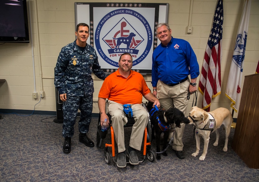 Commander Chadwick Bryant, Naval Consolidated Brig Charleston commander, and Rick Hairston, Canines for Service president, present service dog Malachi (black Labrador) to Rick Hayes, an Air Force veteran, during a ceremony March 21, 2013, at the Naval Consolidated Brig Charleston on Joint Base Charleston – Weapons Station, S.C. CFS is a non-profit health and human services organization that trains service dogs for people with disabilities. Through this program, military prisoners are taught to train service dogs for veterans with disabilities. Since the program's inception, 15 wounded service members have received service dogs. Service dogs are constant companions and can assist veterans with more than 70 tasks, including retrieving and carrying objects, opening doors, and helping with stress and balance difficulties.. (U.S. Air Force photo/ Senior Airman George Goslin)