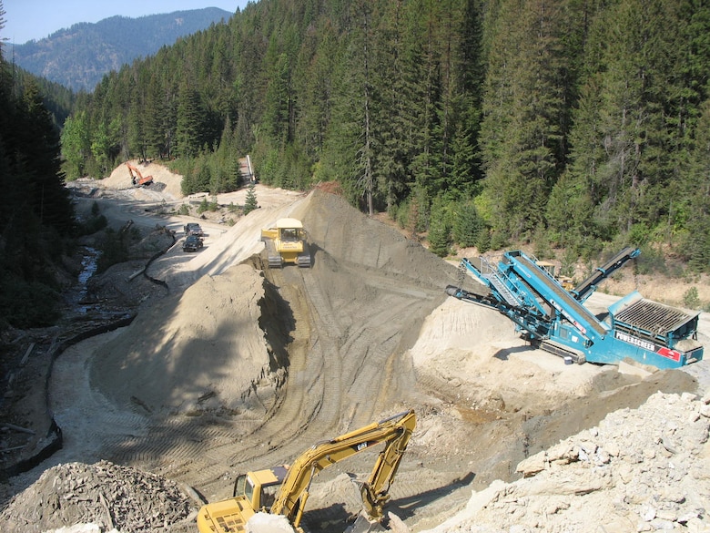 This photo was taken from the vicinity of Tailing Pile 3 looking down stream toward Tailing Pile 1. Tributary Creek is on the left side. The activity in the foreground is the contaminated tailings screening operations used to produce materials for the cushion layer beneath the geomembrane cover systems at the tailings disposal sites. The excavator in the background is working on Tailing Pile 2 excavations. 