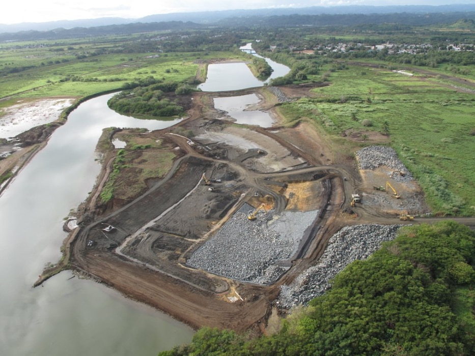 This recent aerial shows construction progress on the Río de la Plata project. The river flows by the municipality of Dorado, about 15 miles west of San Juan on the northern coast of Puerto Rico. Looking upstream, the photo shows channel excavation and placement of stone revetment (right), while the river flows through a temporary diversion channel to the left. The revetment will slow the velocity of storm water coming from the mountains upstream and prevent erosion of the banks. 