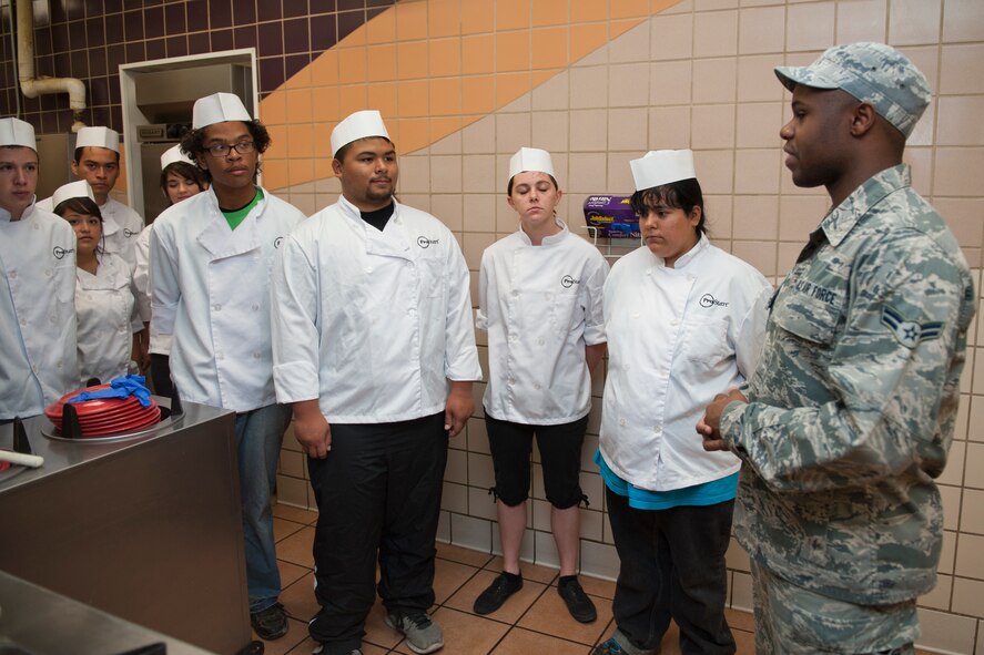 Airman 1st Class Carlton Smith (right), 49th Force Support Squadron culinary specialist, welcomes 18 culinary students from various local high schools to Holloman Air Force Base, N.M., May 8. The students visited the Shifting Sands Dining Facility to learn how the military plans, prepares and serves thousands of meals a day to its personnel. The students were divided into small groups and received hands-on experience in napkin folding, garnishing, serving and cooking. (U.S. Air Force photo by Airman 1st Class Colin Cates/Released) 