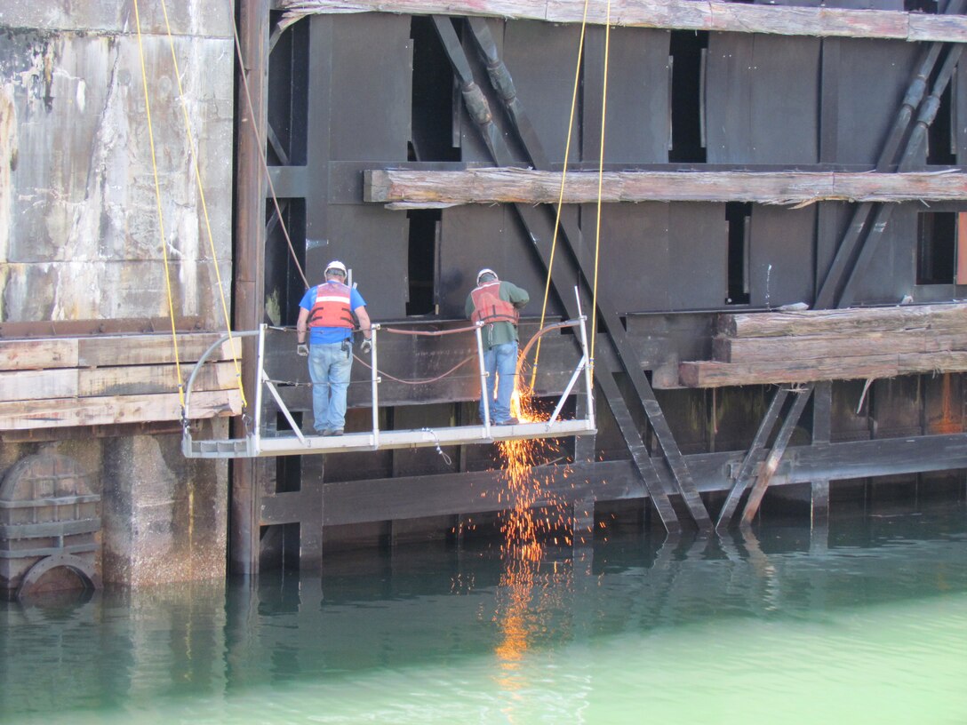 Working on a platform suspended above the water, two workers are busy replacing worn fenders from the lower gates of the MacArthur Lock.  The 12 X 12 inch oak beams line approximately 8 miles of pier walls at the locks and are continually being replaced somewhere on the facility.