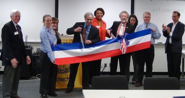 Toastmasters District Governor Mo Hamilton (center, in red) oversees the ribbon cutting at Gribble Gatekeepers chartering ceremony.  Also pictured are (left to right) Toastmasters Division Governor Paul Chamberlain, Erin Rooks, Cynthia Wood, Stuart Davis, Bob Pietrowsky, Lauren Leuck, Jeff Petty and Ken Lichtman. 