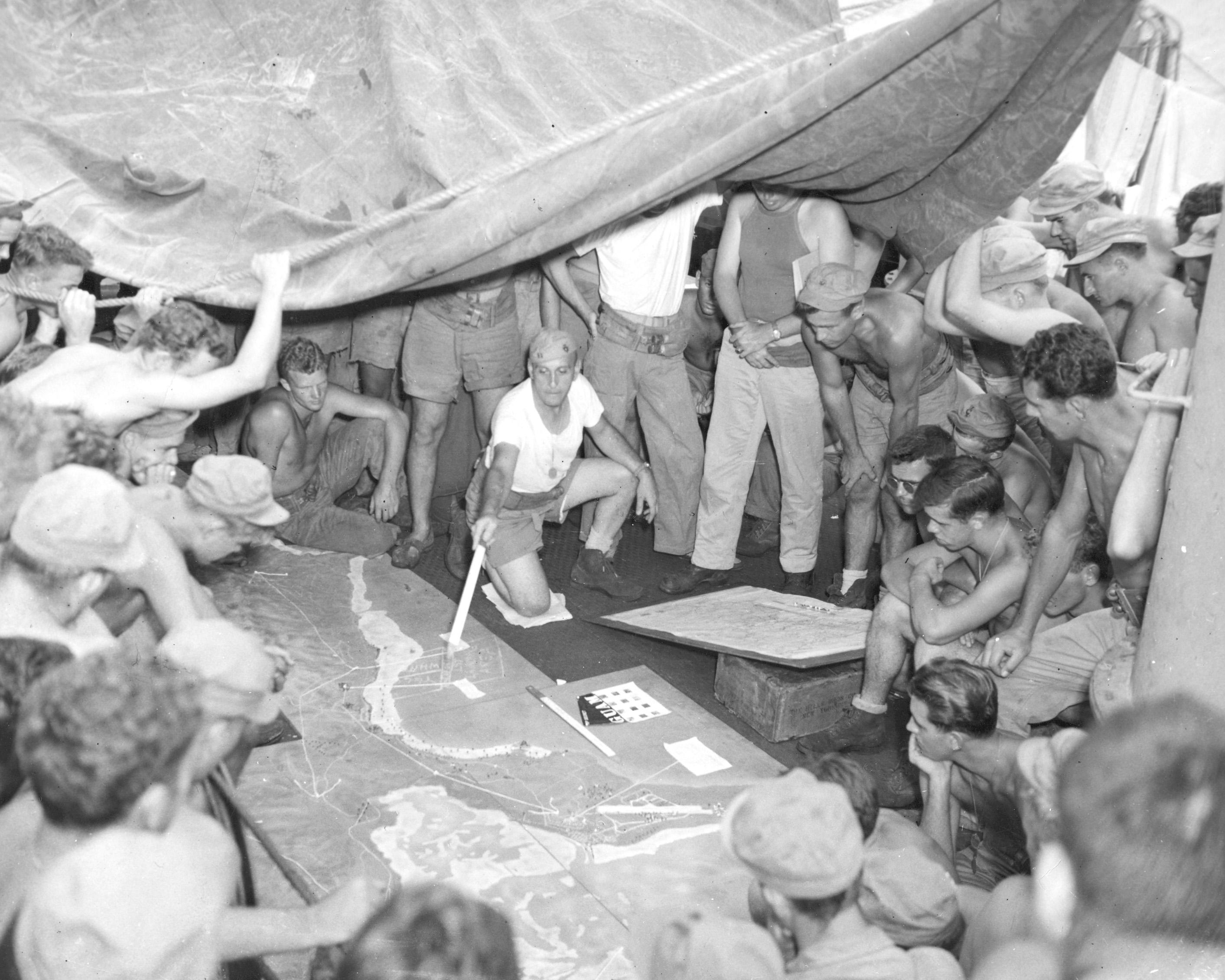 Men of the pack Howitzer Battalion aboard an LST, study a relief map of the area on Guam on which they are to land.  Captain Phillip P. Santon, Battalion Intelligence Officer, is aiding the lecture.  The Guam assault by the Third Marine Division and the First Marine Provisional Brigade came July 20, 1944, after a 17-day aerial and naval bombardment which established a record tonnage of explosives dropped on a Japanese position.  The Marine advance was steady and the former American possession, captured by the Japanese on December 10, 1941, was completely won by August 9, 1944.