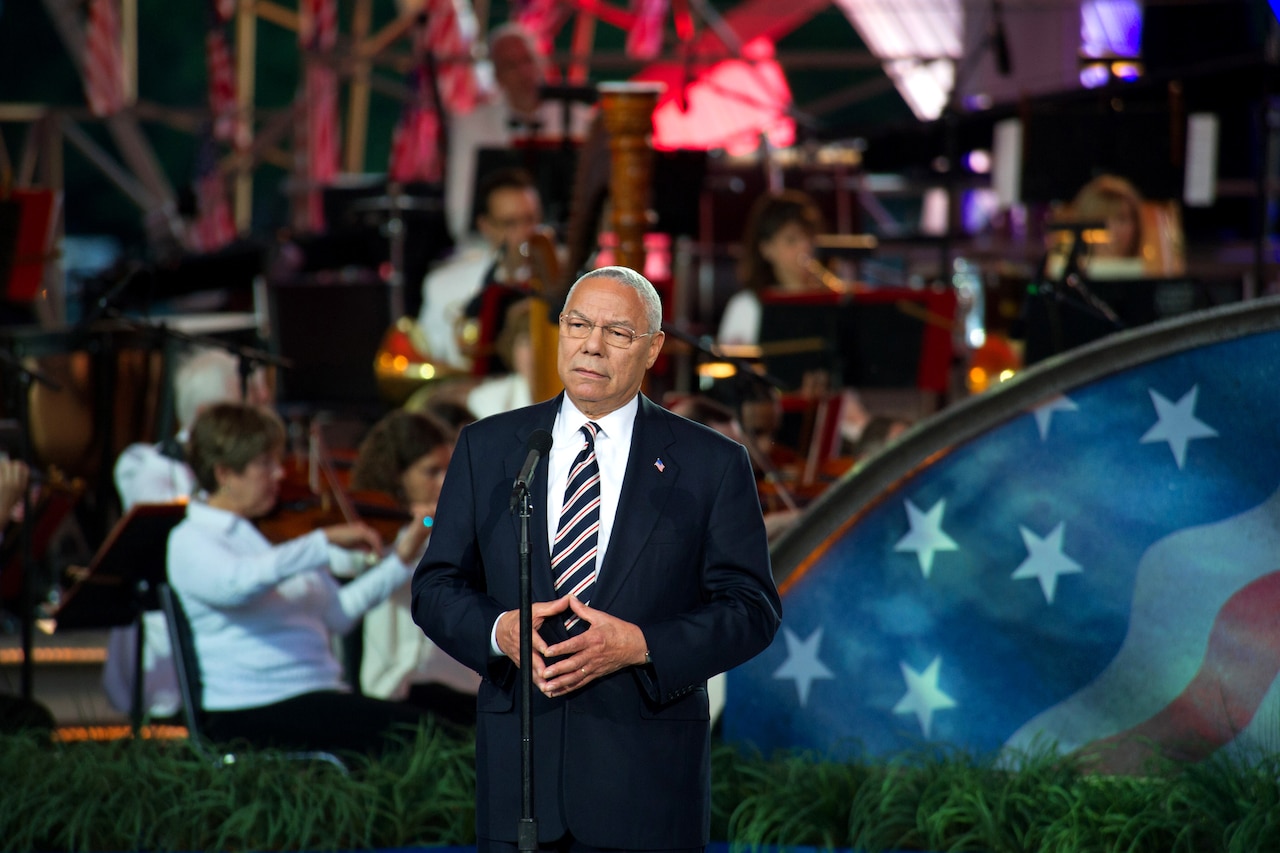 Retired Army Gen. Colin Powell stands at a microphone with concert players on a stage in the background.