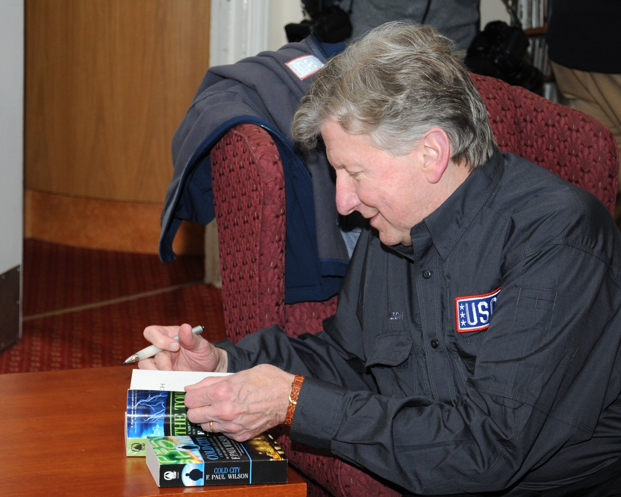 F. Paul Wilson, author, signs a copy of his book for a fan Oct. 31, 2013, at the library on RAF Mildenhall, England. Authors Kathleen Antrim, Phillip Margolin, Harlan Coben, Wilson and Heather Graham took part in a book-signing event as part of the USO tour, Operation Thriller IV. Following the book-signing event, some of the authors returned to the U.S. while others continued on to Brighton, England, for a fantasy writers’ convention. (U.S. Air Force photo by Gina Randall/Released)