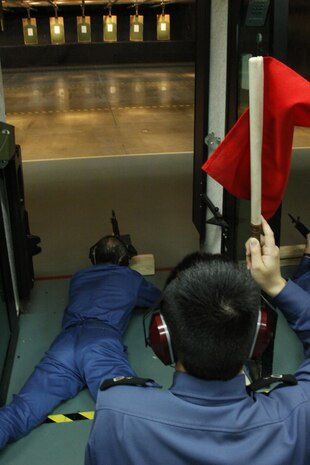 A Japanese Maritime Self-Defense Force sailors snap into the prone position and aim down range while another JMSDF sailor holds a red flag in the air during rifle re-qualification and familiarization training at the Indoor Small Arms Range here Dec. 2. Red flags were used to notify the range coach who still had live rounds.