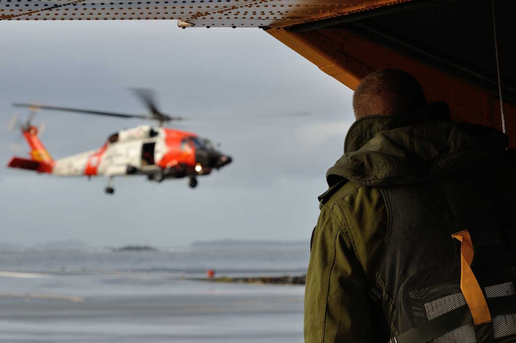 Sgt. Murray Slack, a Royal Canadian Air Force flight engineer on board a 442 Transport and Rescue Squadron Cormorant helicopter, takes a pause from pre-flight checks to watch as the crew of a United States Coast Guard (USCG) H-60 helicopter from USCG Air Station Sitka, conducts hoist training on the morning of Oct. 29, 2013. The 442 Squadron crew was stopping over in Sitka on their way to a joint Canada/U.S. Arctic Search and Rescue Exercise (SAREX) at Joint Base Elmendorf-Richardson, Alaska. Cross-border SAREXs are conducted regularly to ensure both Canadian and American SAR personnel are familiar with each others' operations in the event of an emergency response involving crews from both the Canadian Armed Forces and U.S. military. (Image by Capt Trevor Reid, 19 Wing Public Affairs)