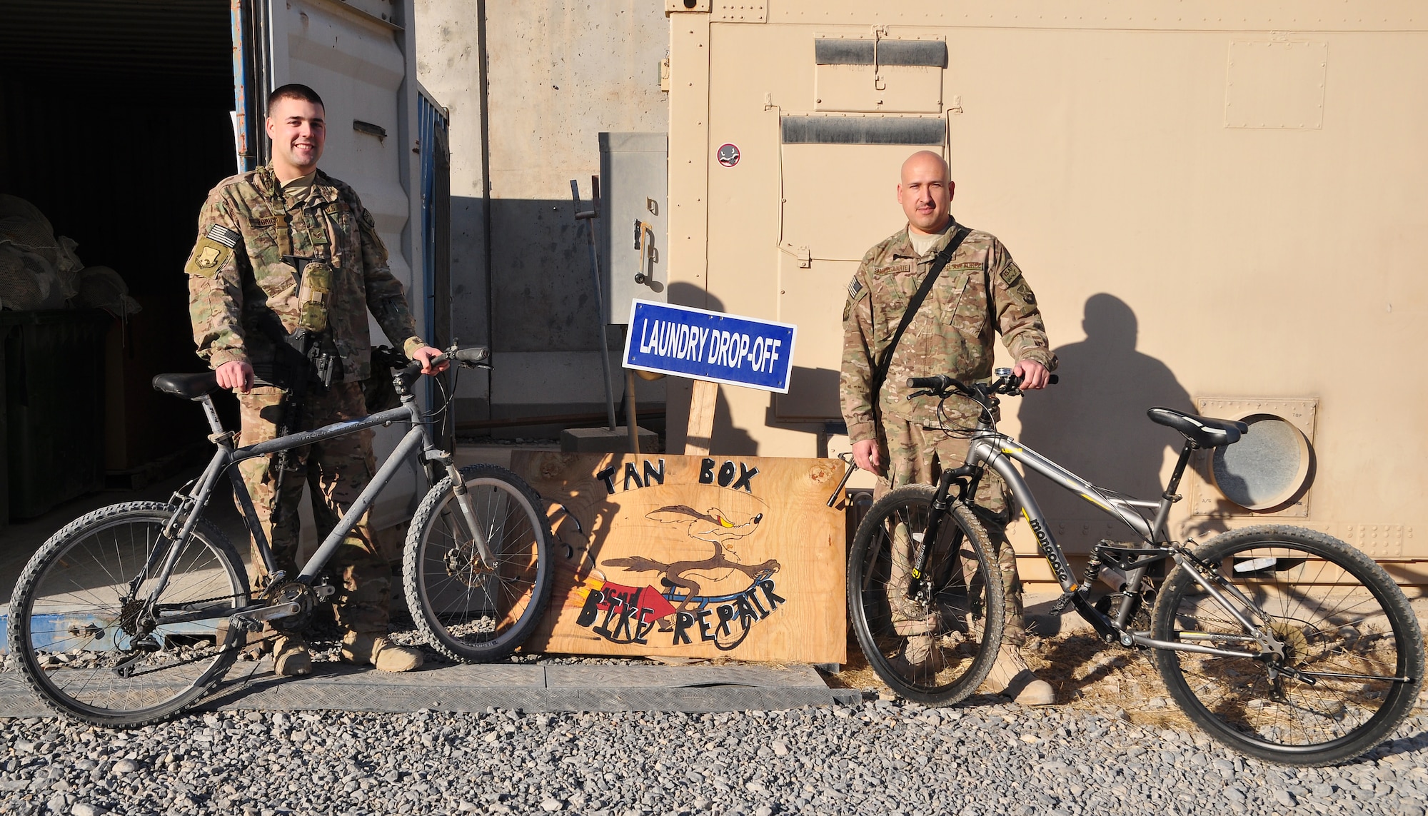 Tech. Sgt. Juan Sanchezduarte (right) and Senior Airman Mark Zorich (left), both assigned to the 451st Expeditionary Logistics Readiness Squadron, pose in front of the plywood “Tan Box Bike Repair” sign at Camp Losano, Kandahar Airfield, Afghanistan, Nov. 11, 2013. Service members at KAF can get free bicycle repairs from Tan Box. (U.S. Air Force photo by Capt. Jason Smith)