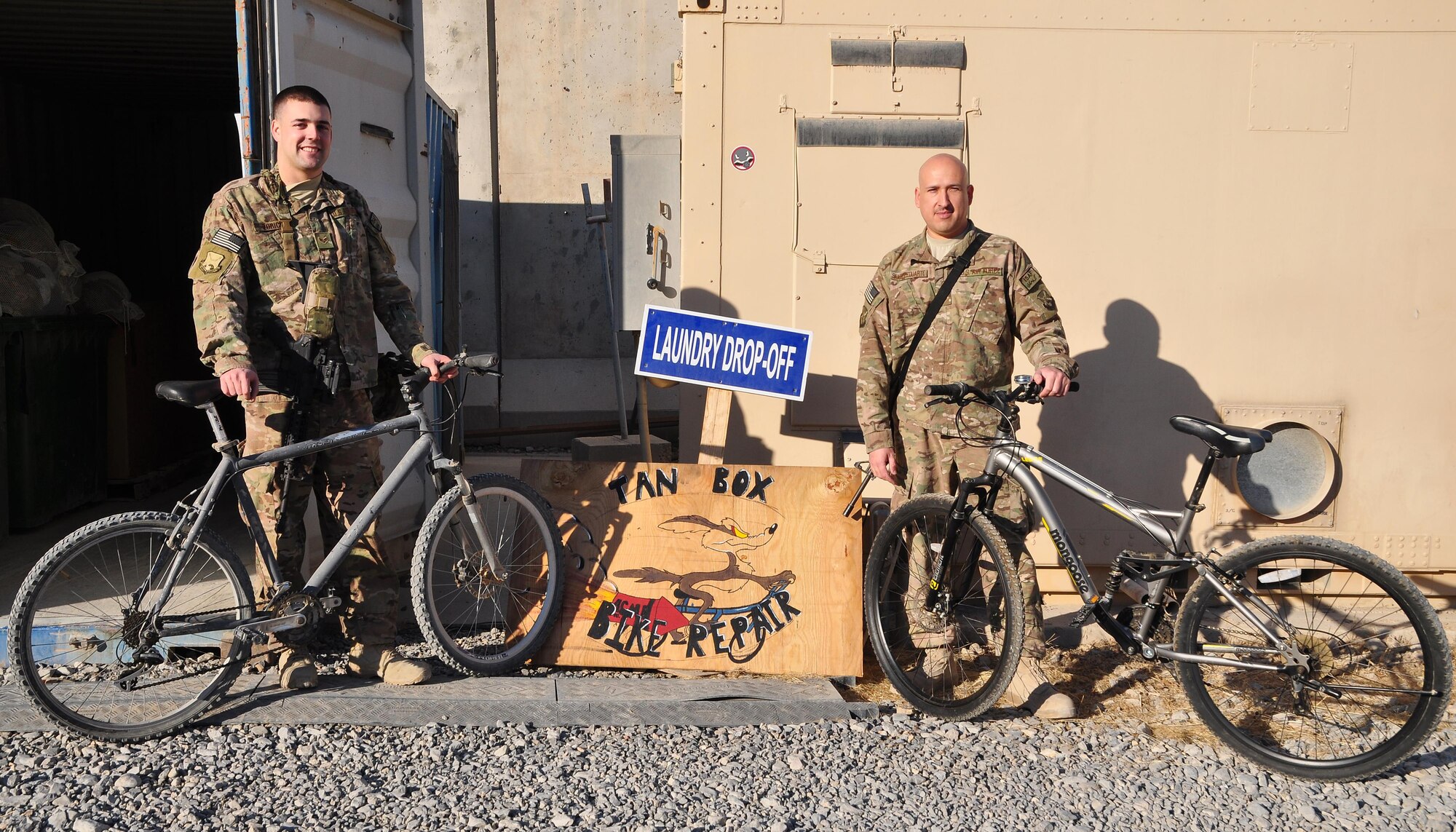 Tech. Sgt. Juan Sanchezduarte (right) and Senior Airman Mark Zorich (left), pose in front of the plywood “Tan Box Bike Repair” sign Nov. 11, 2013, at Camp Losano, Kandahar Airfield, Afghanistan. Service members at KAF can get free bicycle repairs from Tan Box. Both are assigned to the 451st Expeditionary Logistics Readiness Squadron.