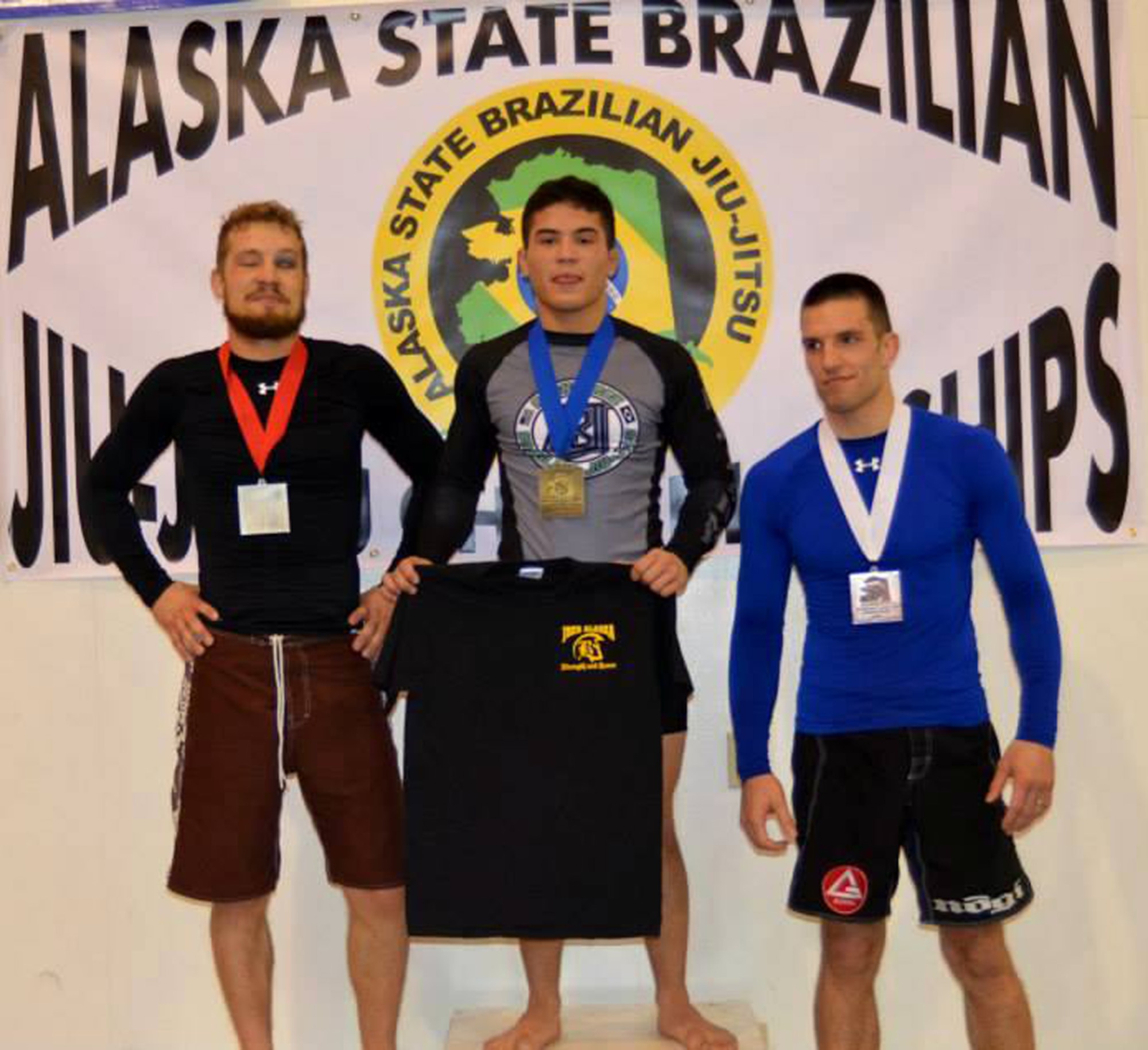 Pfc. Jason Hayden, a paratrooper with B Company, 725th Brigade Support Battalion, displays his unit shirt as he stands atop a podium after winning in the Alaska State Jiu-Jitsu Championship tournament Nov. 2 hosted at the Lumen Christie High School in Anchorage. Hayden won the state championship in the 149 pound and under category and in the overall “Absolute” category. (Courtesy Photo)