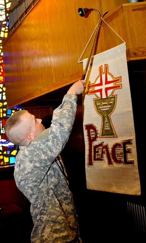 U.S. Army Spc. Stephen Maynus, Fort Eustis chaplain assistant, hangs a new Ecumenical Colors in the Regimental Chapel at Fort Eustis, Va., in preparation for the Cathloic Mass, Sept. 30, 2013. Maynus said even though he does not wear the cross on his uniform like his chaplain, he shares the same pride in serving others. (U.S. Air Force photo by Staff Sgt. Wesley Farnsworth/Released)