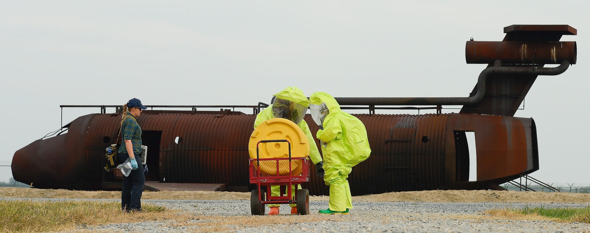 Jessica Feil of Alliance Solutions Group,( left) monitors the progress of  Staff Sgt. Jaimee Scott (middle), 436th Aeromedical Squadron and Technical Sgt. Justin Alexander, 43th Civil Engineer Squadron, as they move toward a simulated contamination site at Dover Air Force Base, Del. on Oct. 3, 2013. The airmen are participating in a unit training exercise while being evaluated by Feil. In the background is an aircraft fire-training device used by the Dover Fire Department. The exercise tested the bases ability to respond to a natural disaster and resulting contamination to water supplies. (U.S. Air Force photo/Greg L. Davis)