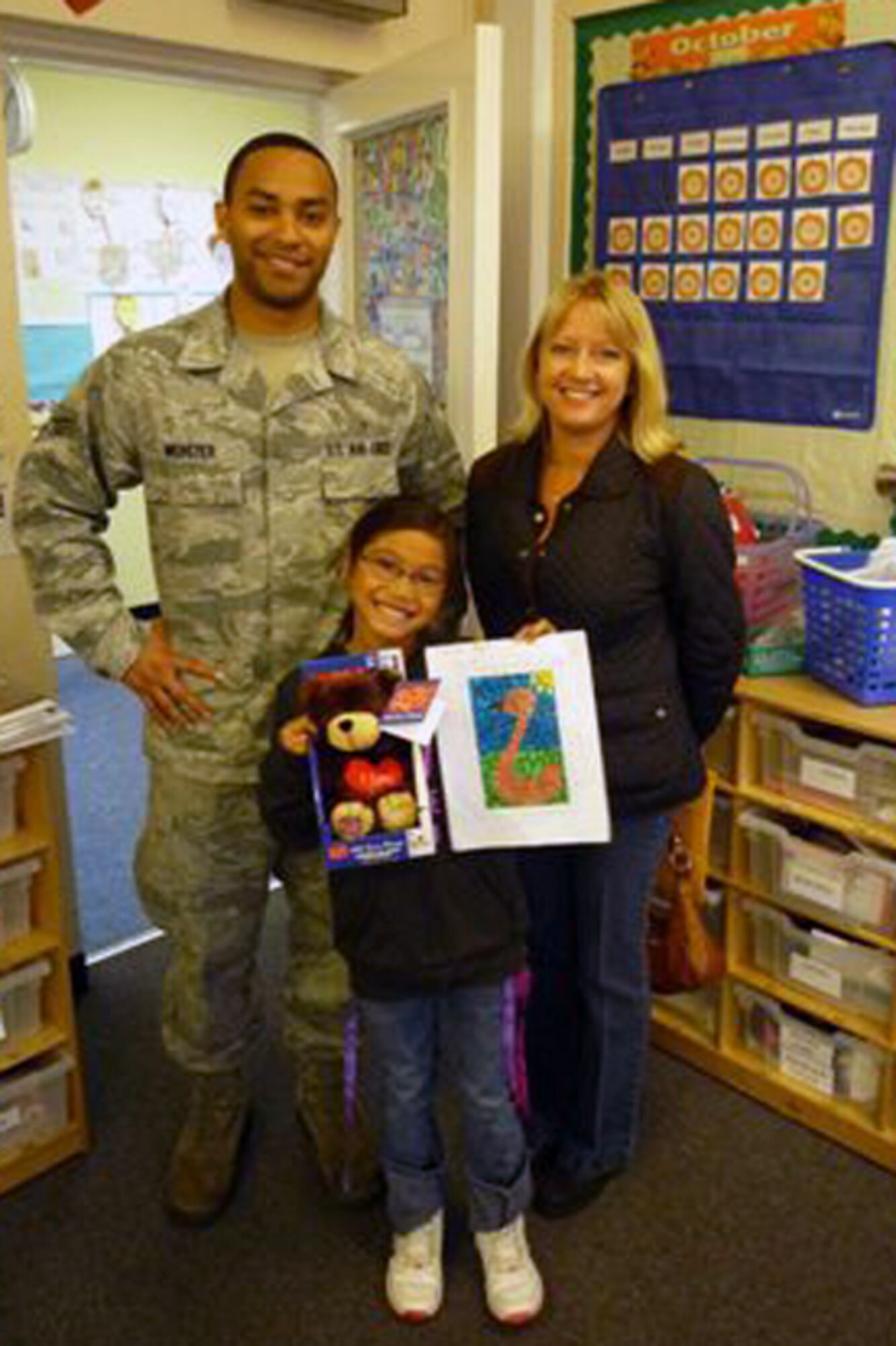 RAF ALCONBURY, United Kingdom – Senior Airman Jarron Webster and Linda Paine, both from 423rd Communications Squadron Alconbury Post Office, present Mariah Camacho with an In the Mail Box "I Love You" Teddy Bear after she was announced as one of nine winners of the 2013 October National Stamp Collecting Month coloring contest the Alconbury Post Office coordinated at the Alconbury Elementary School. (Photo courtesy of Dr. Terry Emerson)